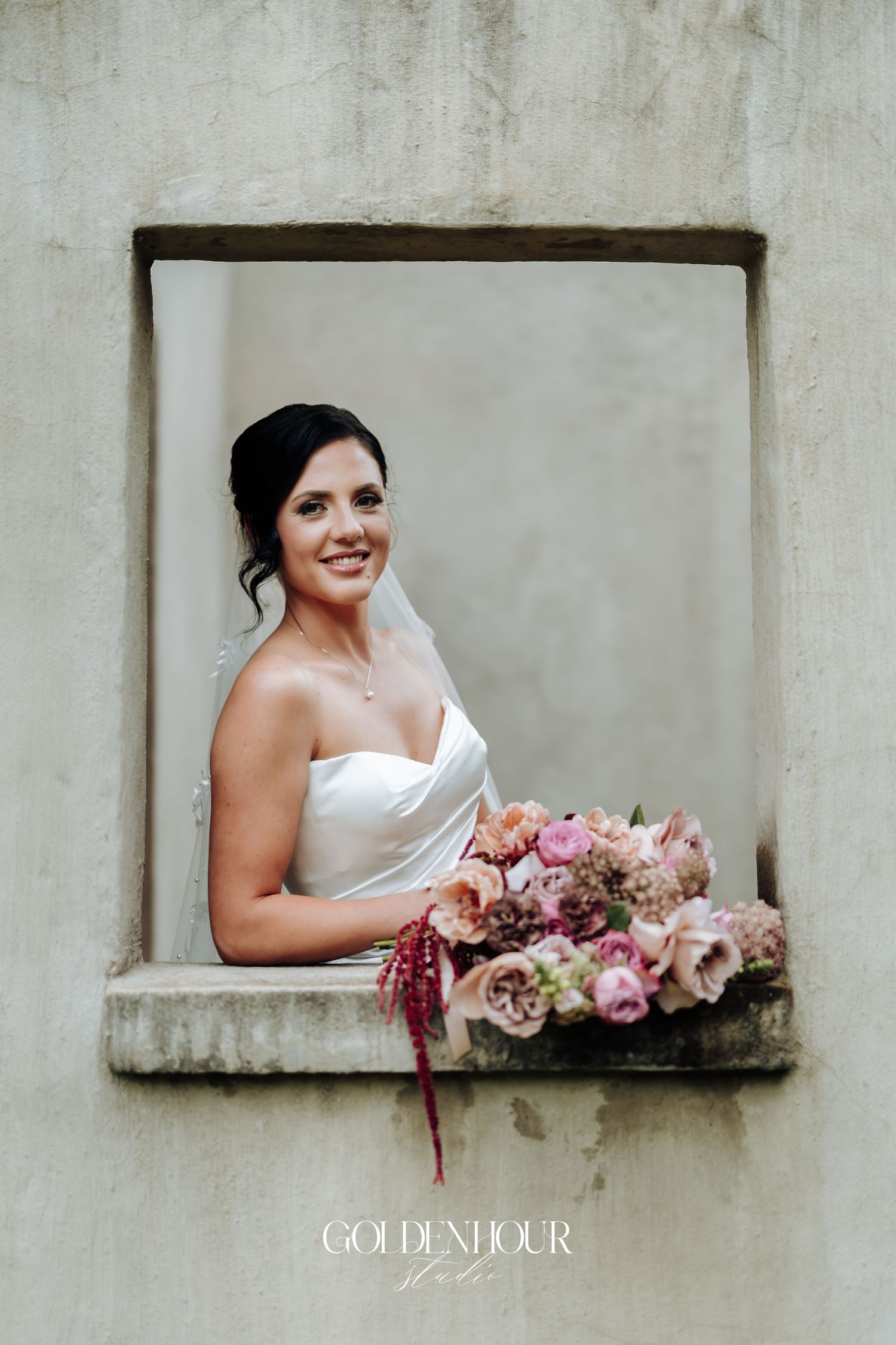 Smiling bride in strapless white dress holding pink bouquet, posing in stone window frame outdoors.