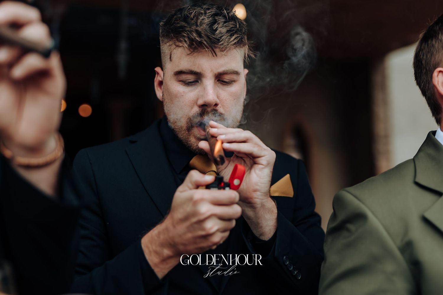Groomsman in black suit lights a cigar with a red lighter at a wedding celebration.