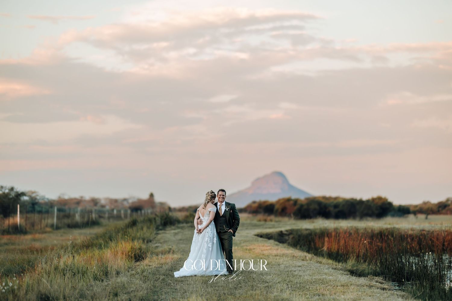 Bride and groom standing on a scenic path at sunset with a mountain silhouette in the background.