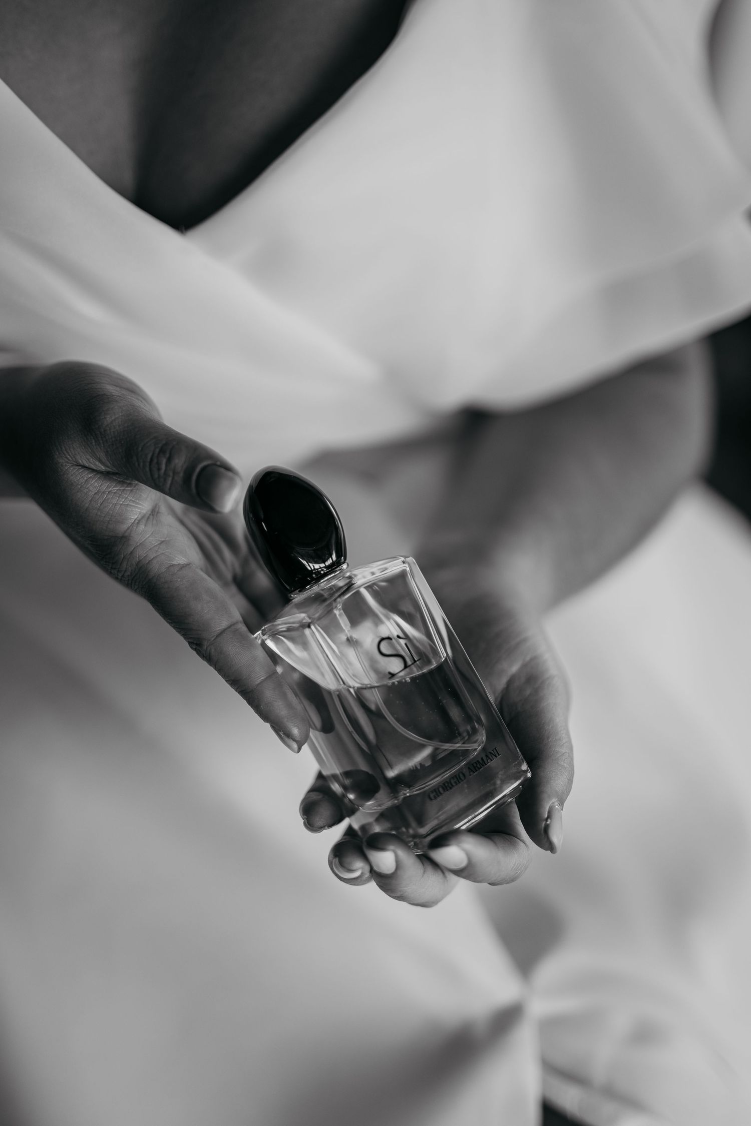 Black and white photo of hands holding Giorgio Armani Si perfume bottle, wearing white robe.