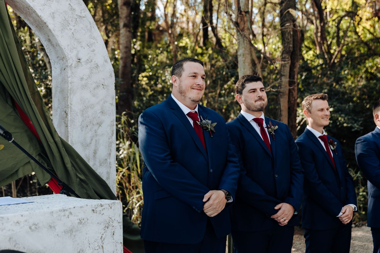 Three groomsmen in navy suits with burgundy boutonnieres stand at an outdoor woodland wedding ceremony.