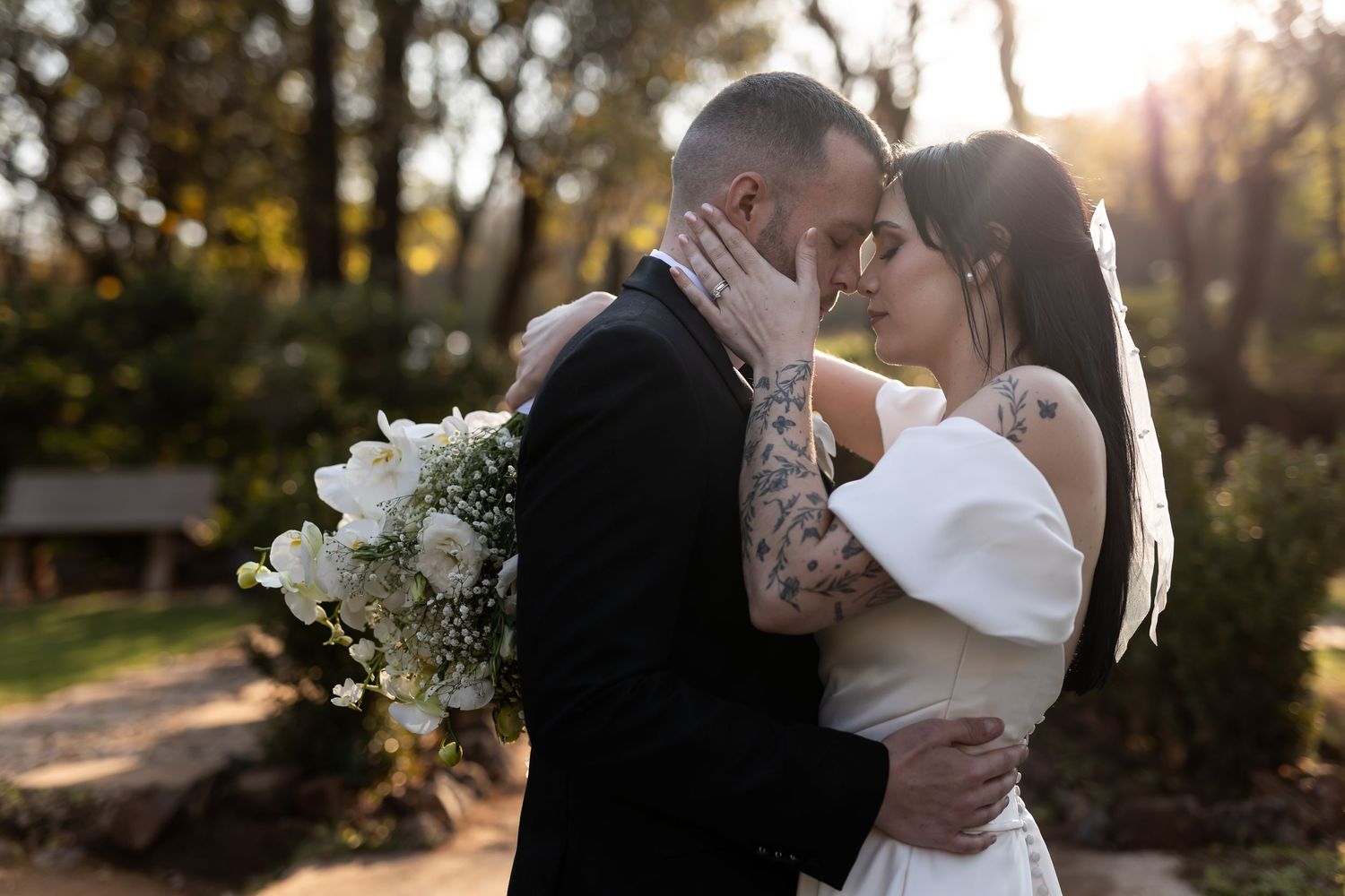 A romantic wedding couple embraces outdoors at sunset while holding a white floral bouquet.
