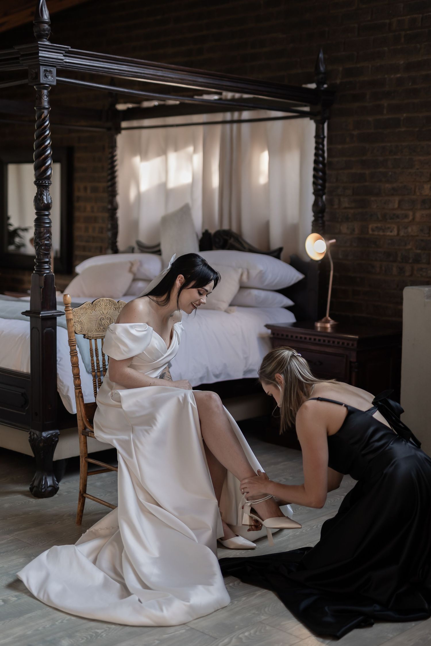Woman in white dress kneeling on floor near rustic dark column and bridal accessories in intimate indoor setting.