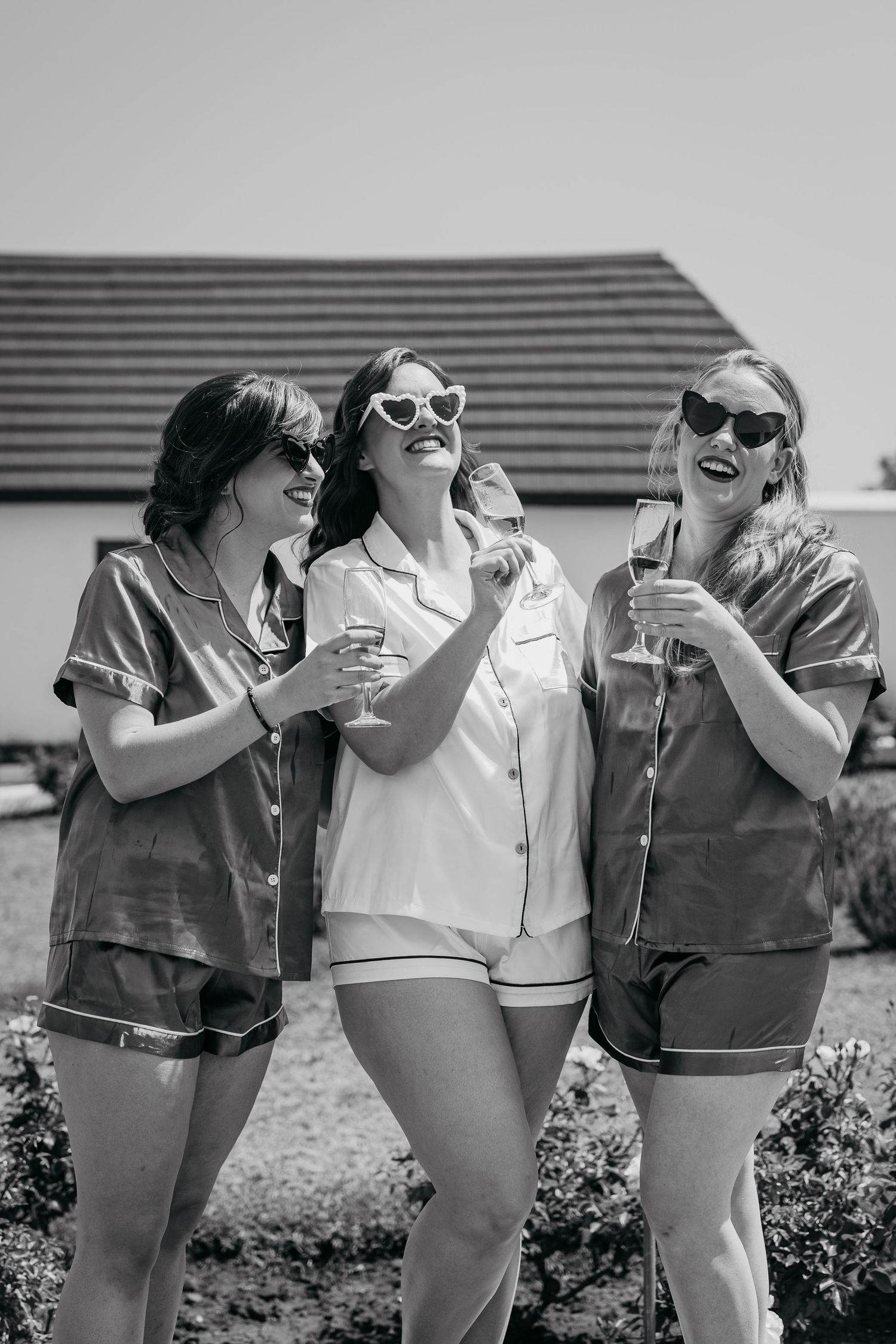 Three friends in sunglasses laughing and having drinks together outdoors on a sunny summer day.