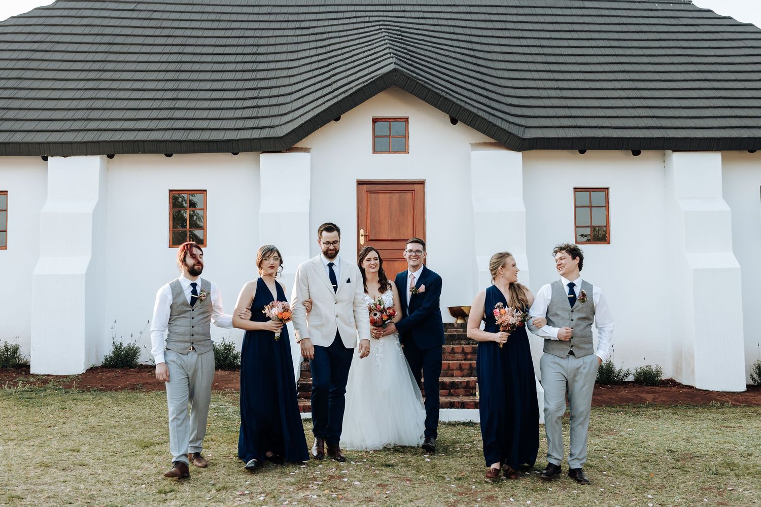 Wedding party in formal attire poses against white wall in panoramic group photo series.
