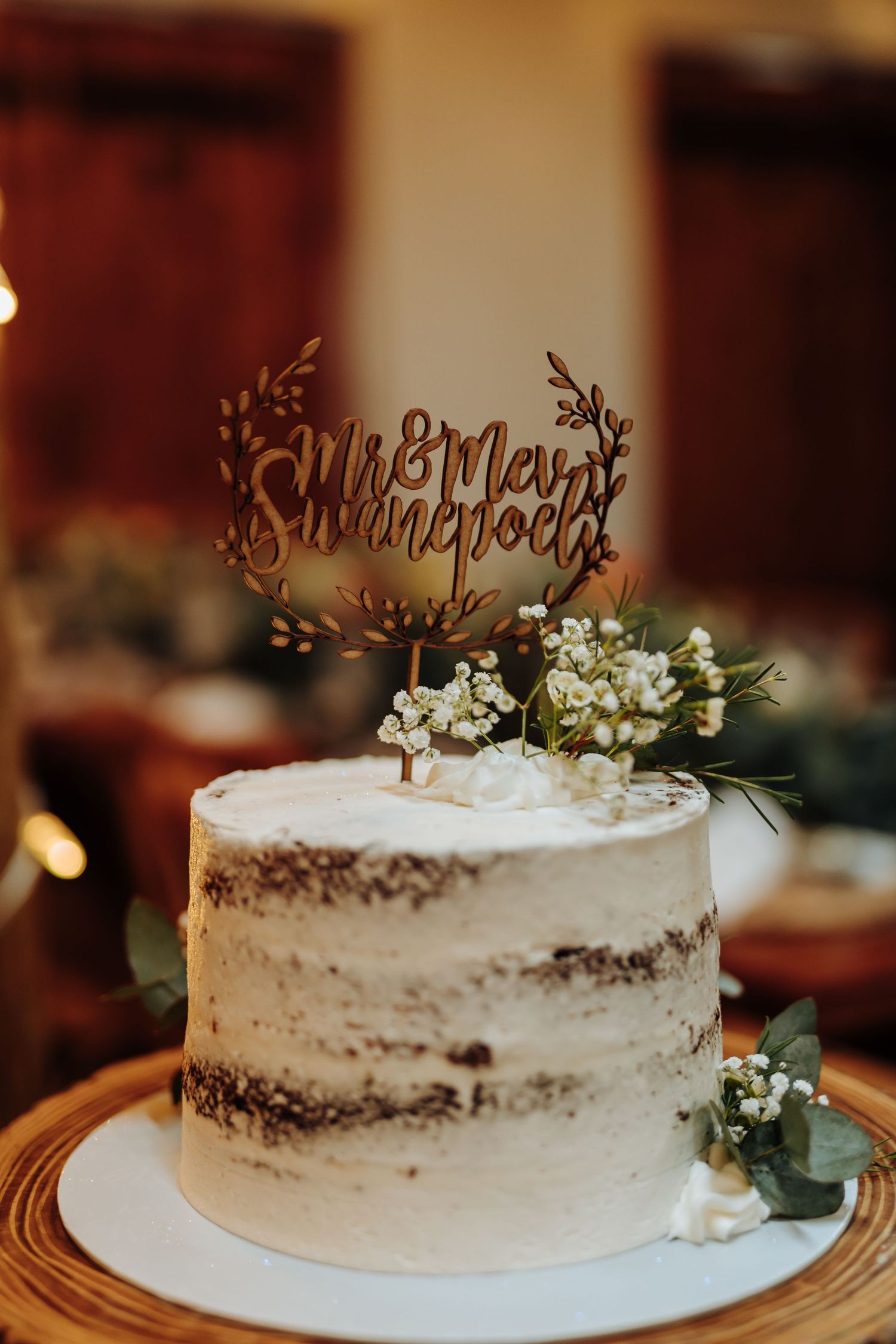 A rustic semi-naked wedding cake decorated with white flowers and a gold 'Mr & Mrs' cake topper on a wooden stand.