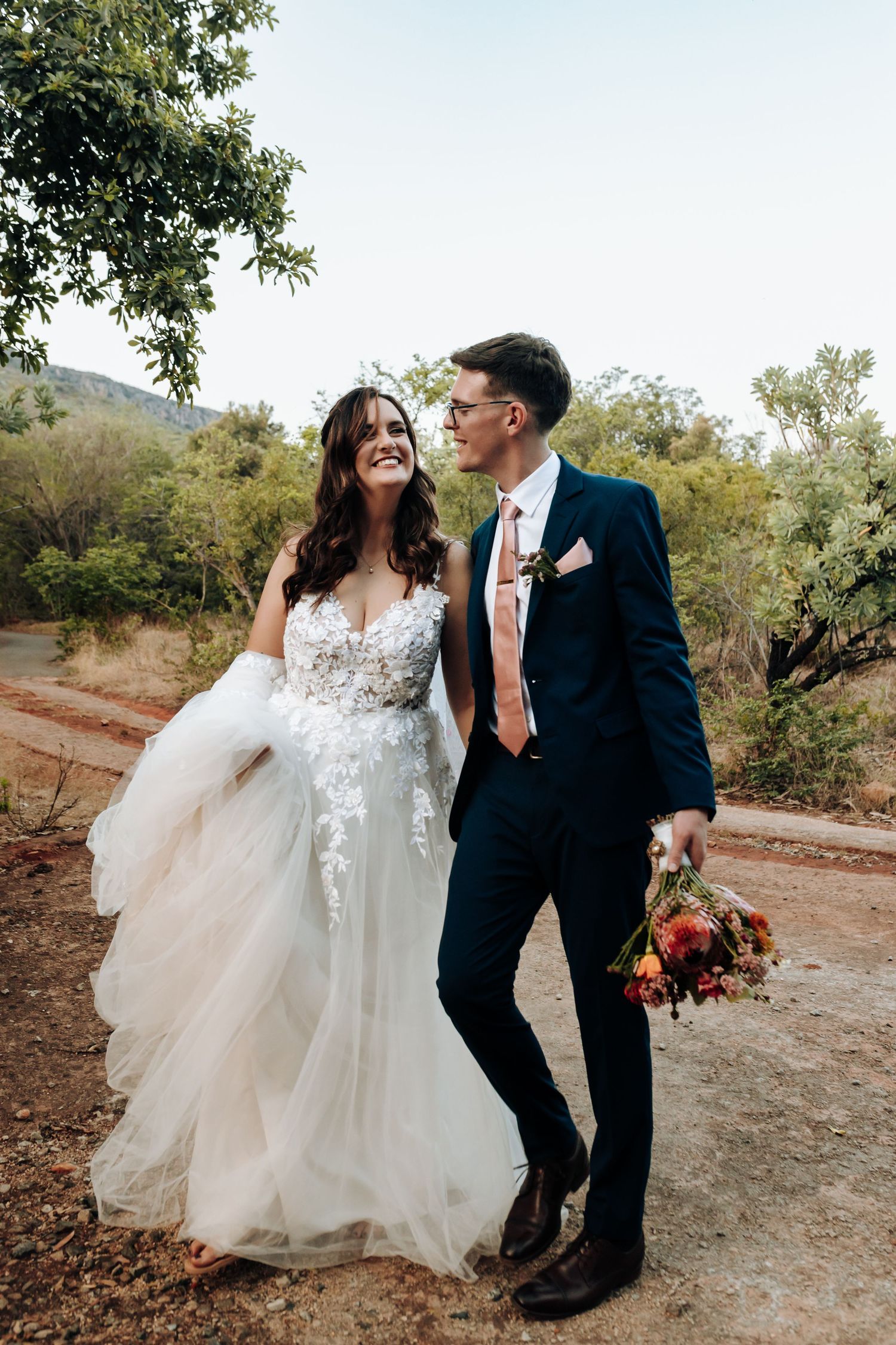 A couple shares joyful moments in their wedding attire against a natural outdoor backdrop with trees and dirt paths.