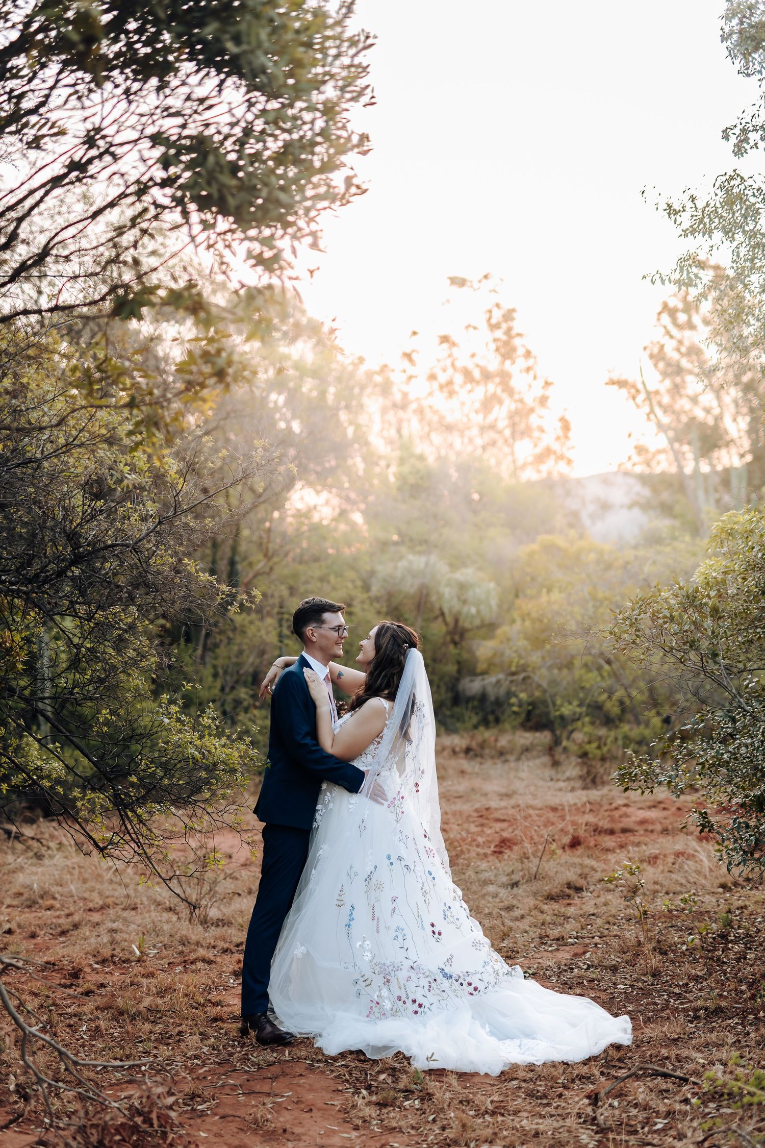 A couple in wedding attire sits together on rustic ground during golden hour as sunlight filters through trees.