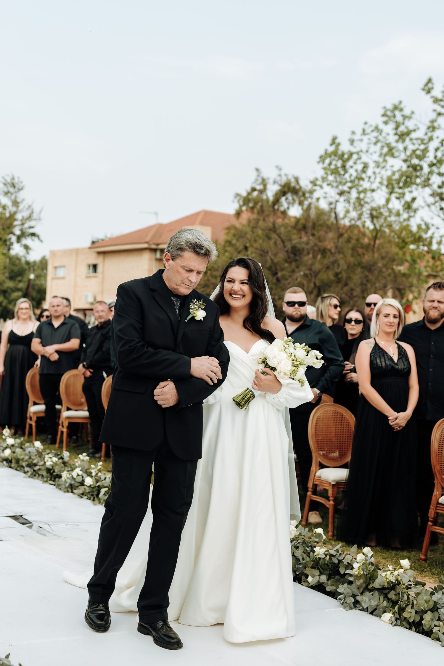 Wedding guests in black attire seated in wooden chairs at an outdoor ceremony with floral arrangements.