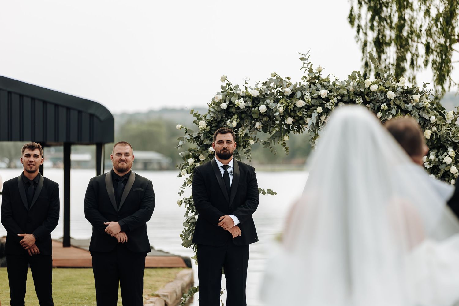 A wedding ceremony takes place outdoors with groomsmen standing at the altar while viewing the bride in her white veil.