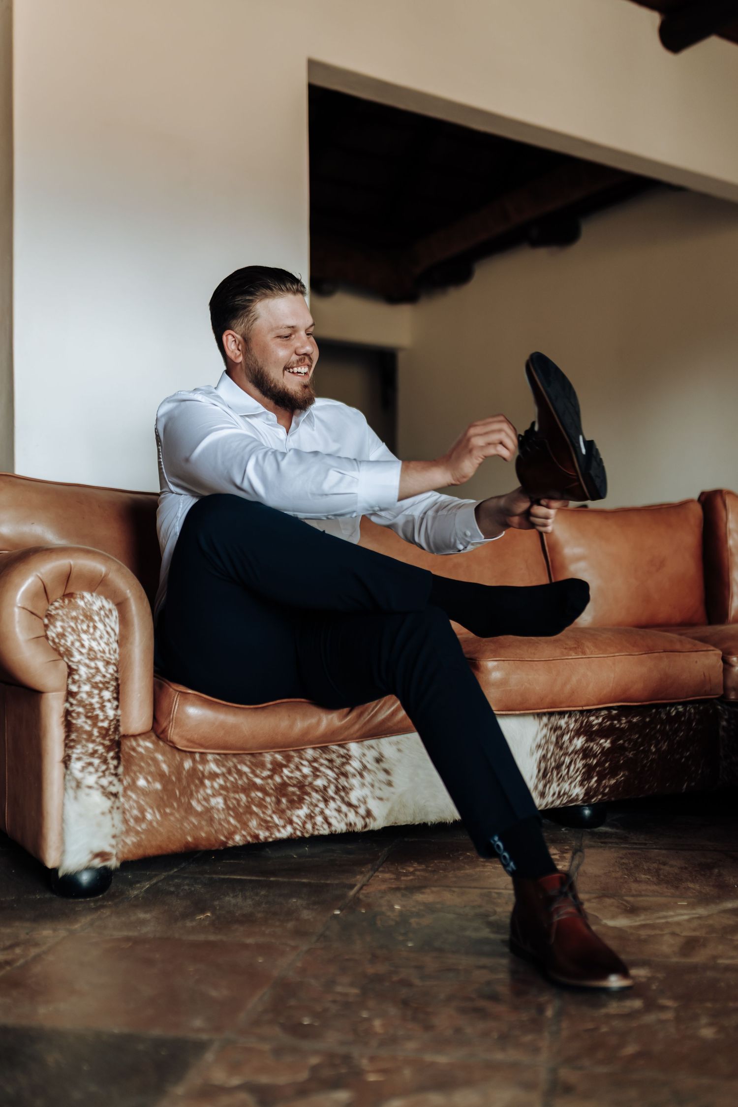 A person in formal attire lounges on a brown leather couch in an indoor setting with dark floors and walls.