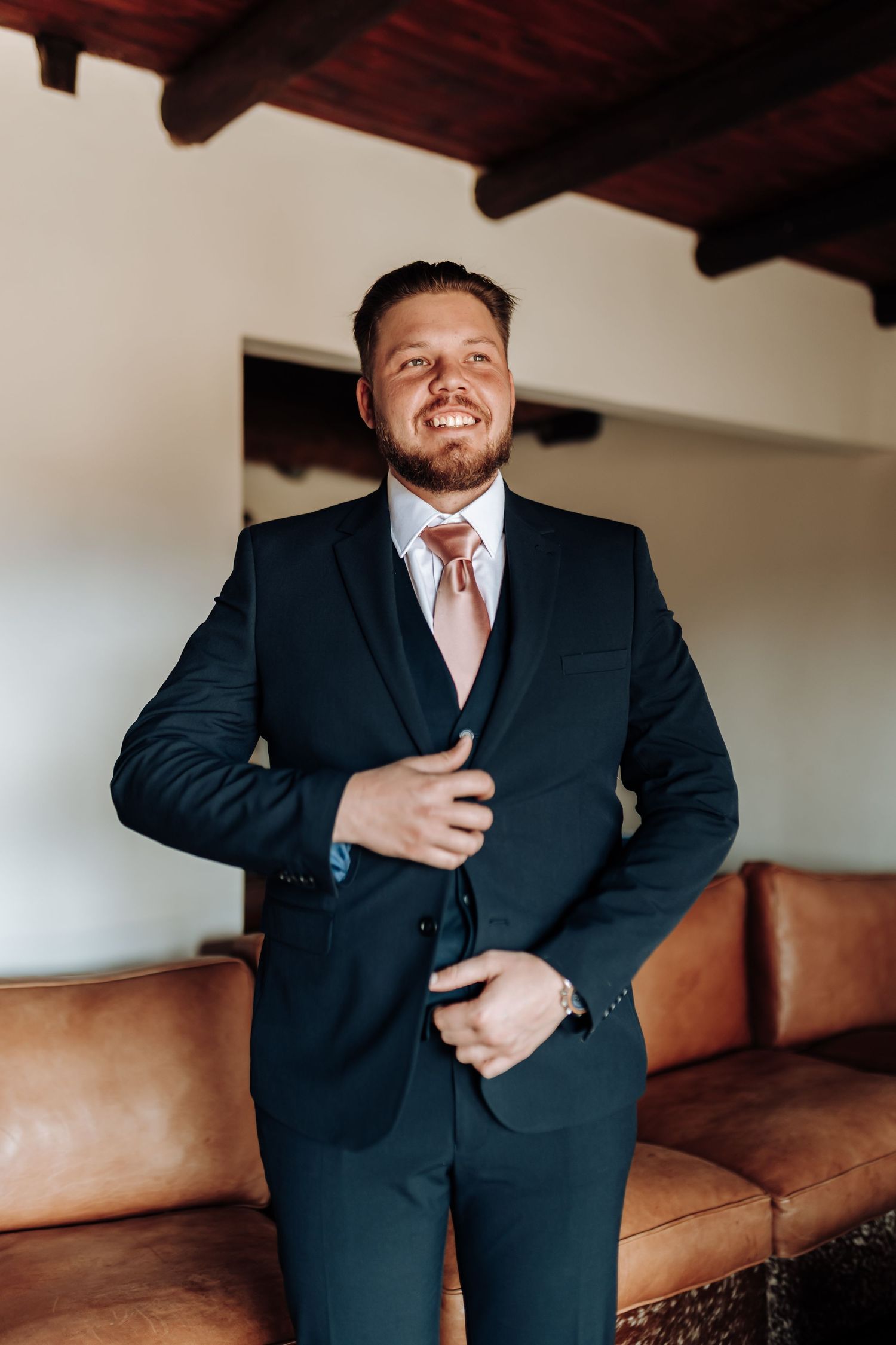 Well-dressed gentleman in a black suit and pink tie adjusts his jacket while standing by leather couch indoors.