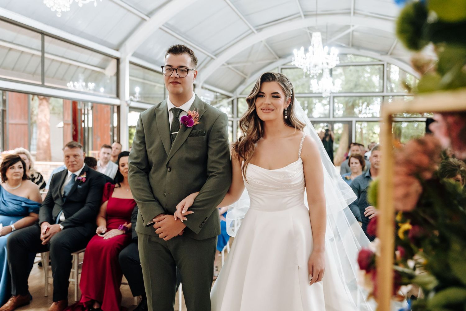 Wedding ceremony in bright sunlit venue with arched glass ceiling and elegant crystal chandeliers.