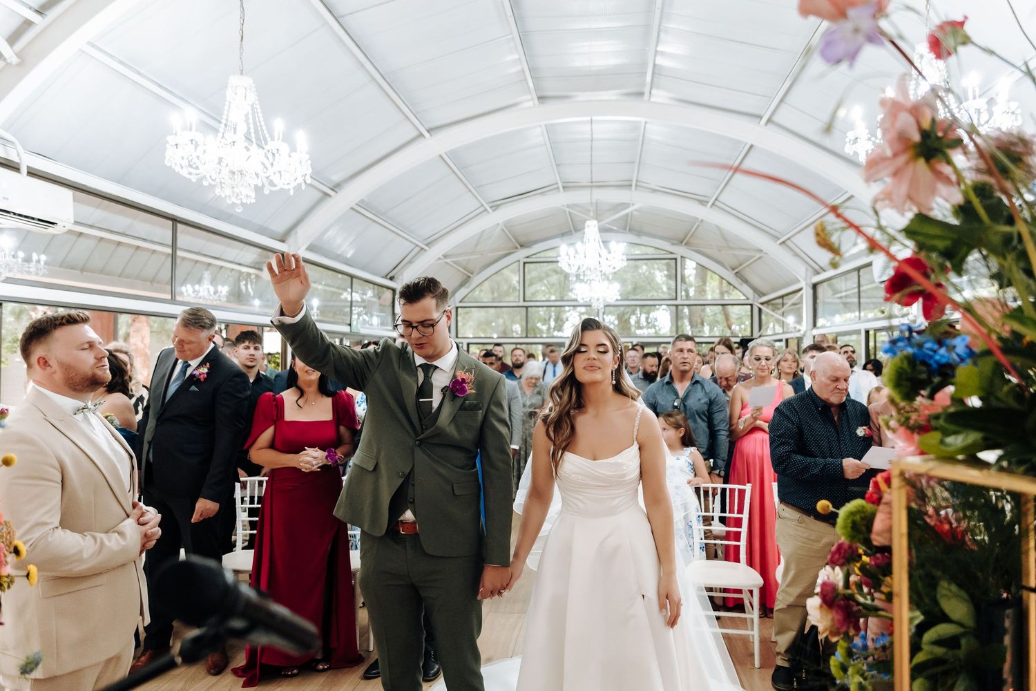Newlyweds celebrate with guests while walking down the aisle in a greenhouse wedding venue with floral decor.
