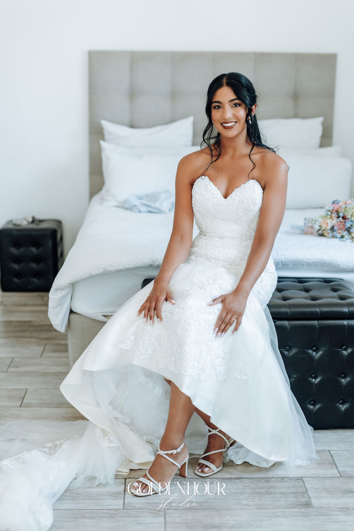 Bride in white lace wedding dress shows off her elegant shoes while sitting on white furniture.