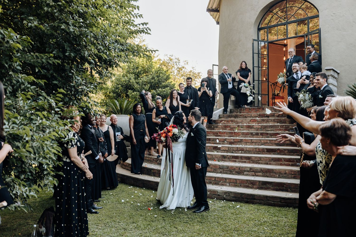 Wedding guests gather outside a white church building with arched doorways during a celebration.
