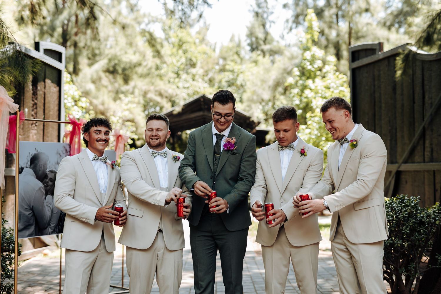 Groomsmen in tan suits and dark suit toast with drinks at an outdoor wedding celebration.