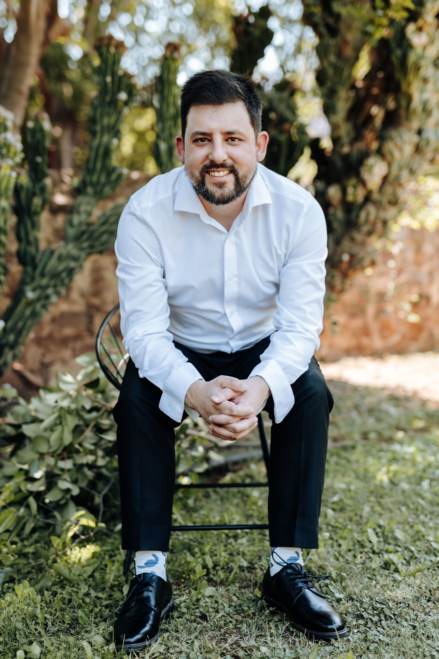 A person in a white dress shirt and dark pants sits casually on grass with trees in the background.