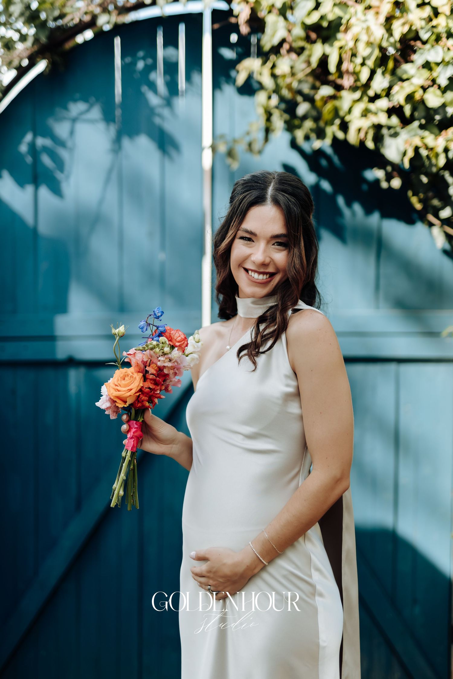 A person in a sleeveless white dress holds a vibrant bouquet of flowers against a blue wall with sunlit greenery.
