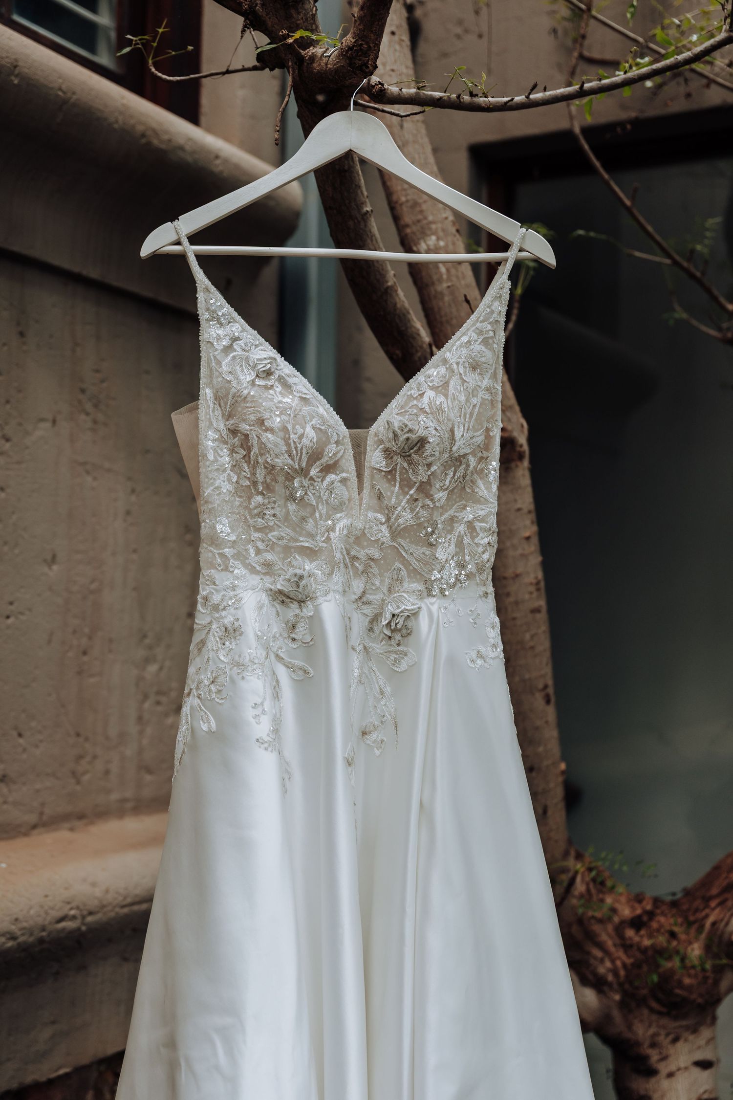 White A-line wedding dress with floral lace bodice and V-neckline hanging on a white hanger from a tree branch.