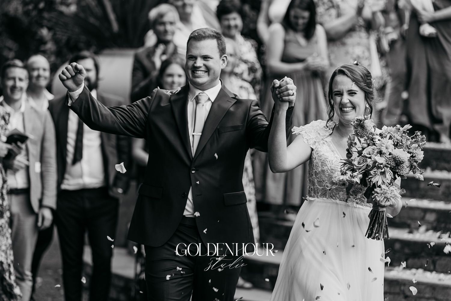 Black and white photo of newlyweds celebrating with raised hands amid confetti.