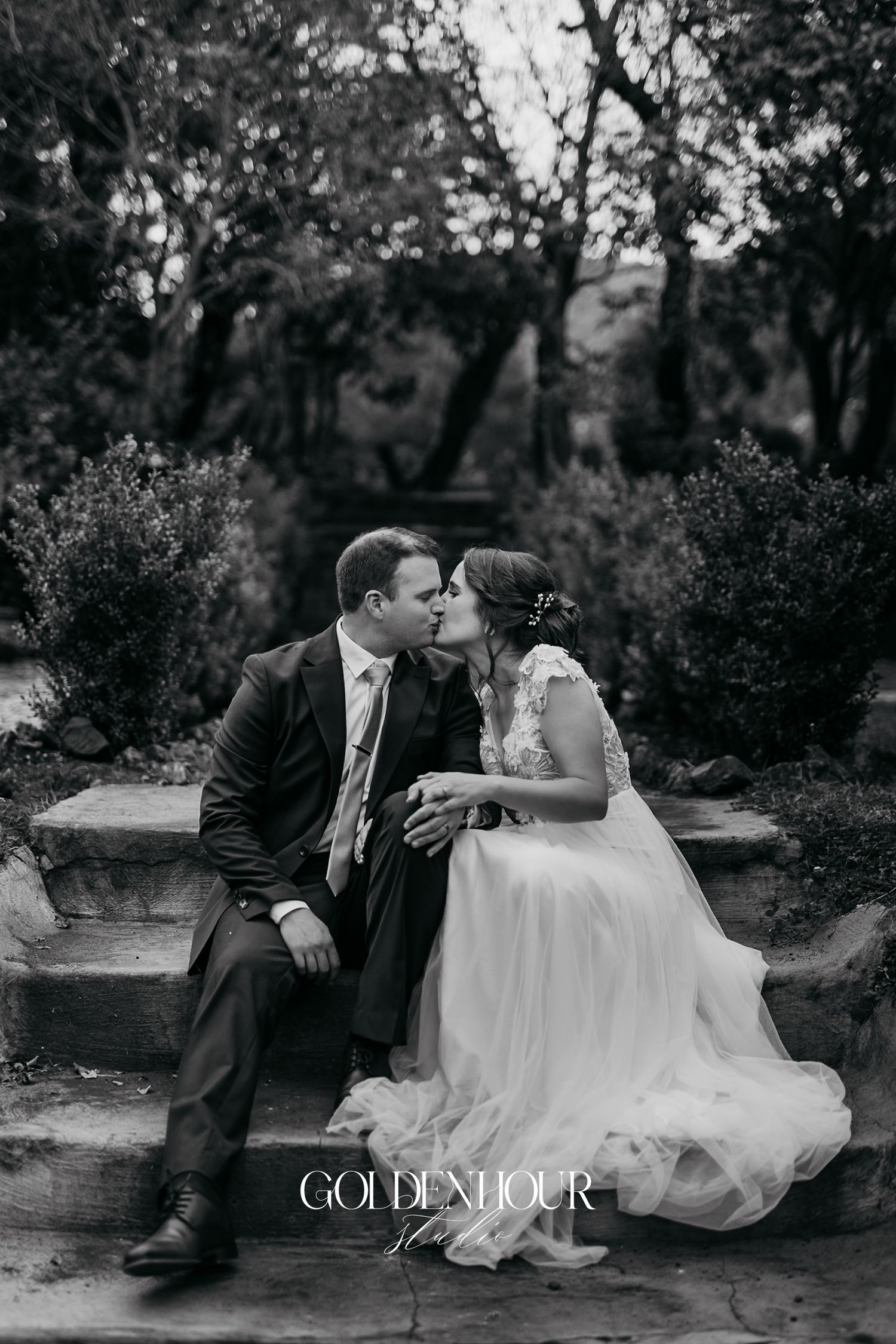 Black and white photo of bride and groom sharing a kiss on stone steps in garden.