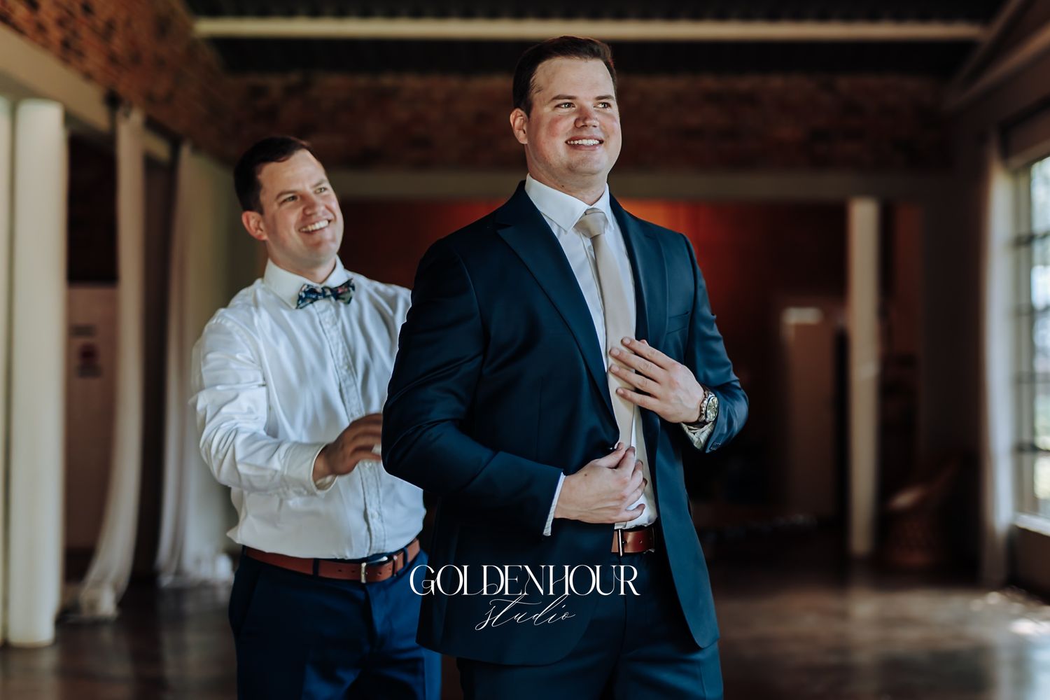 Groomsman helping groom adjust tie in navy suit inside elegant wedding venue.