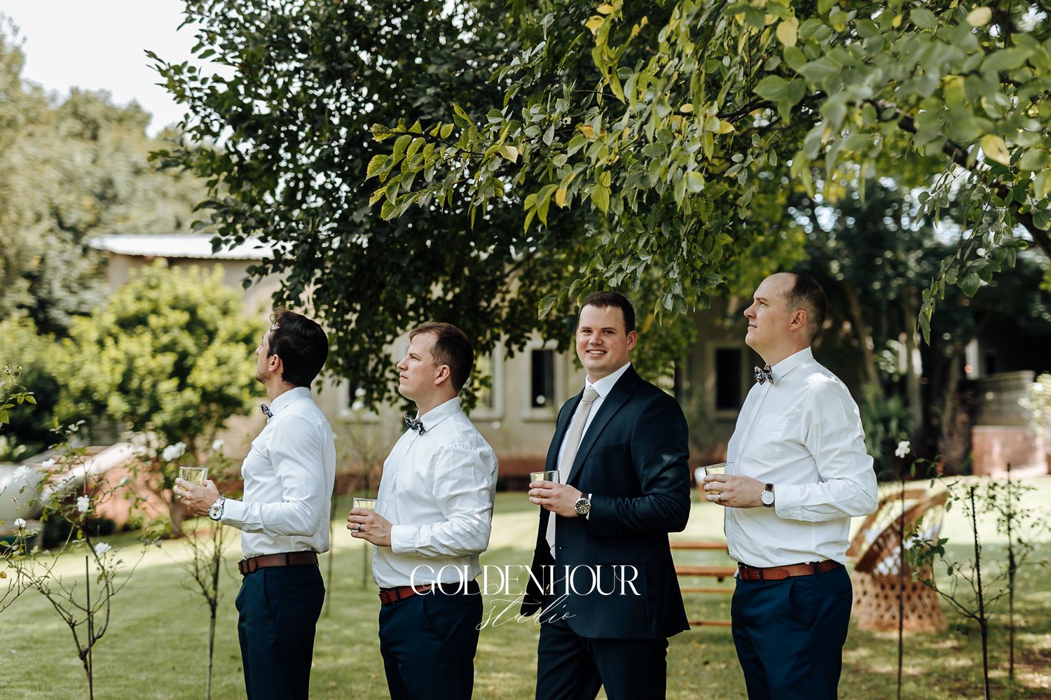 Groom and groomsmen in navy suits relaxing outdoors under a tree at a wedding.