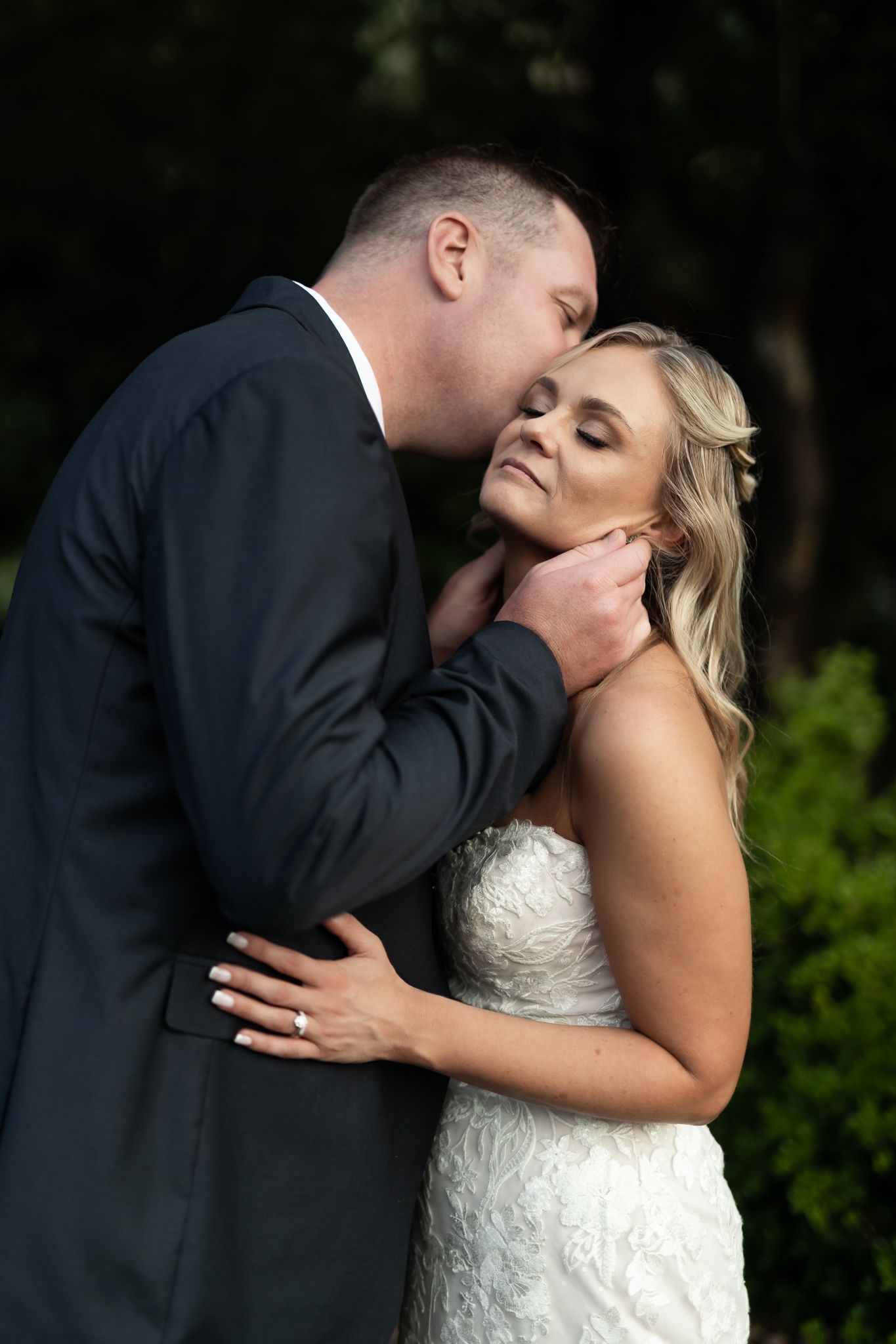 An intimate moment between newlyweds in formal attire against a dark natural backdrop.