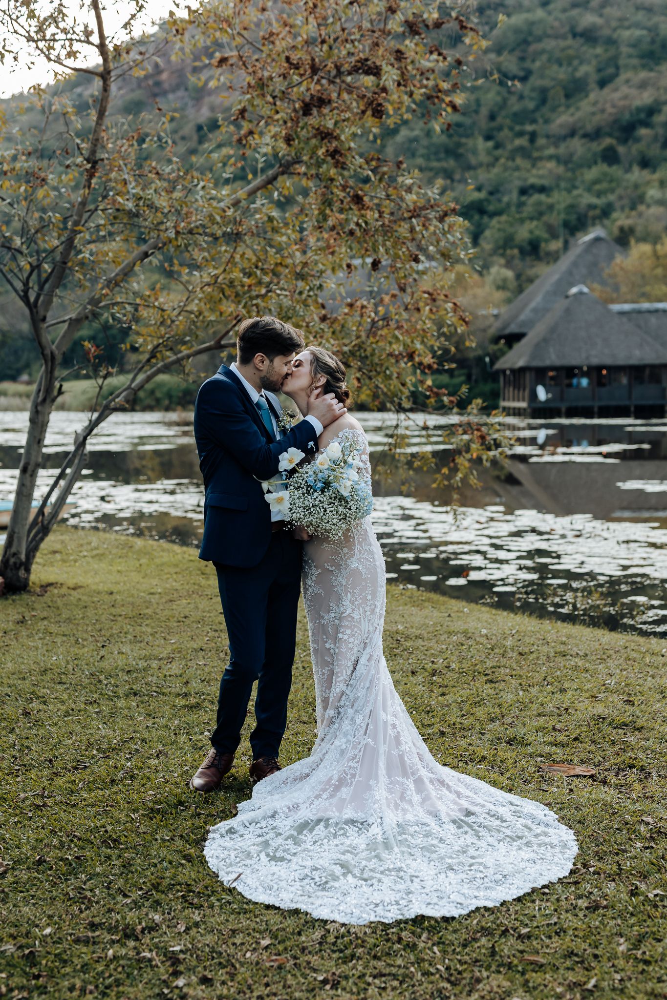 A wedding portrait by a lake showcasing the bride's long lace train and scenic mountain backdrop.