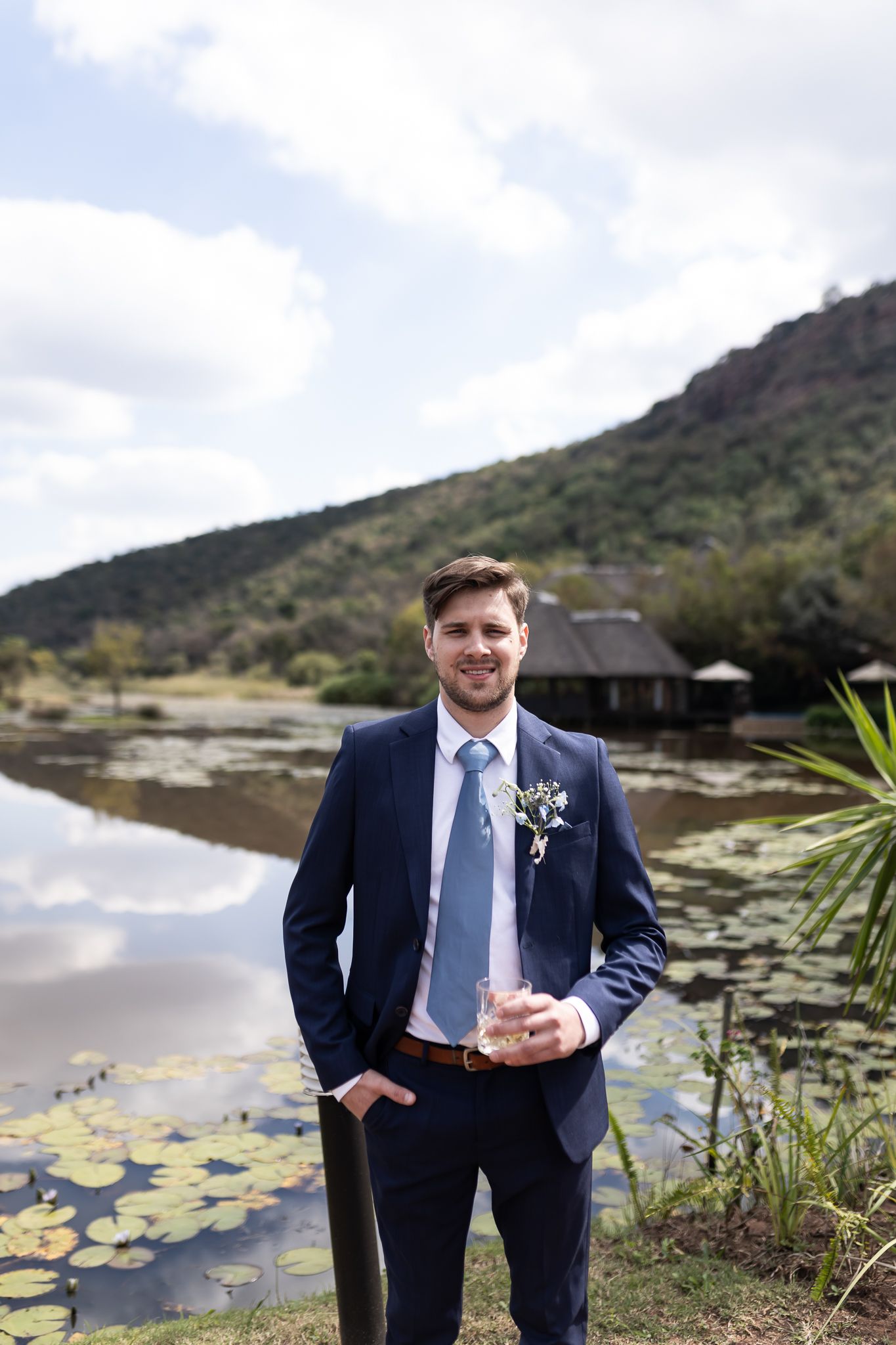 Groom in navy suit standing by scenic lily pond with mountains in background.