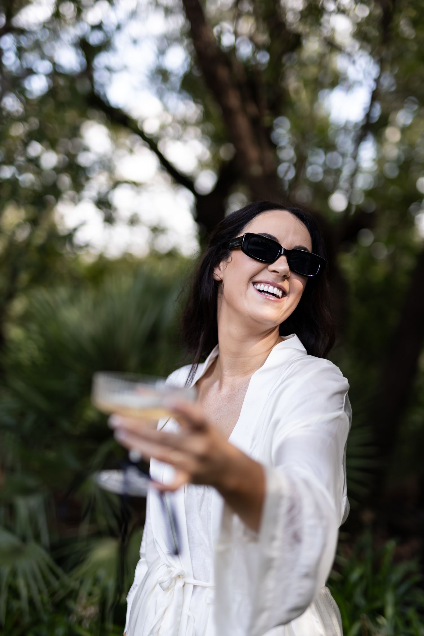 Person in white robe and sunglasses laughs while holding champagne glass in garden setting.