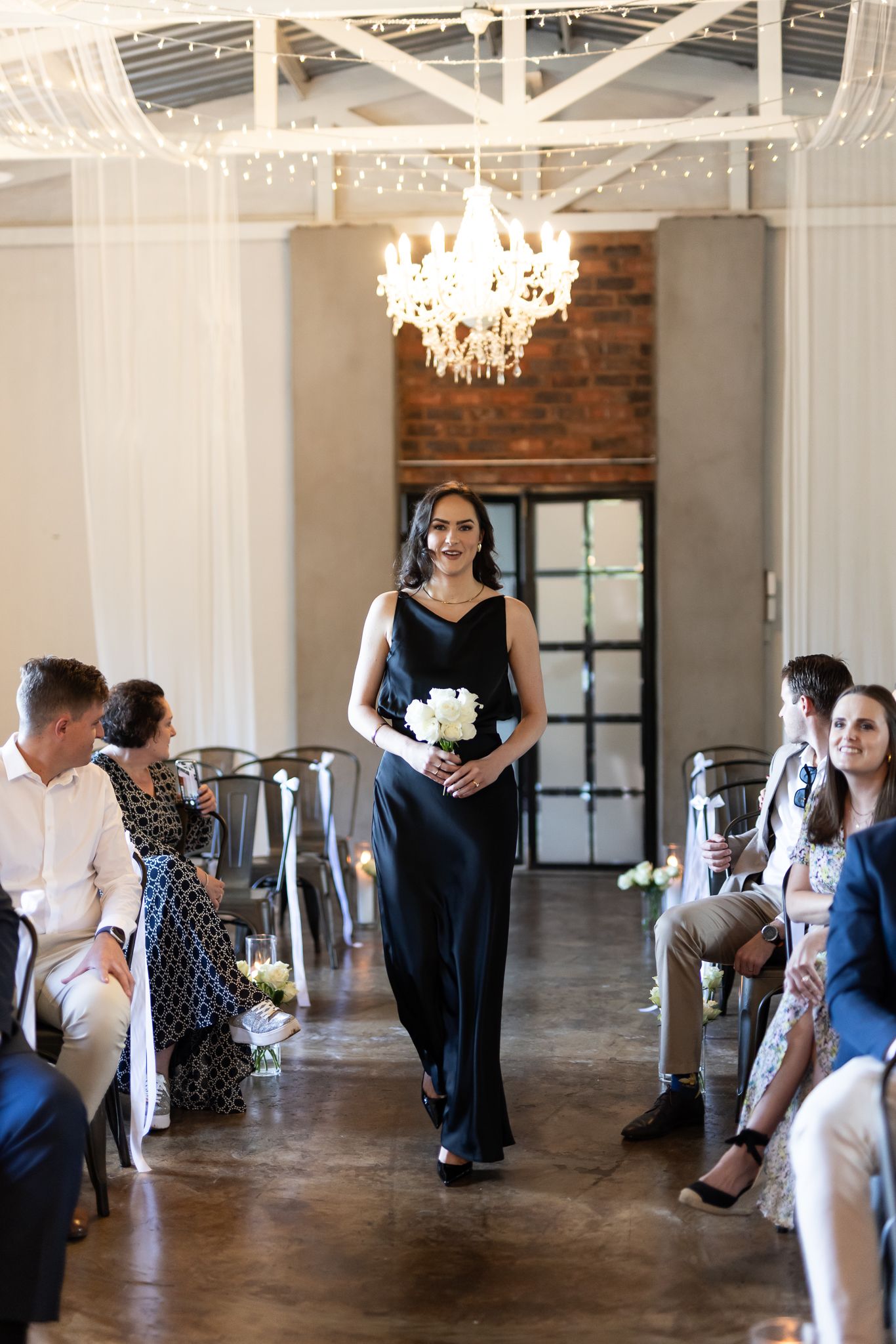 Bridesmaid in black gown processes down aisle with white bouquet in modern venue with exposed brick.