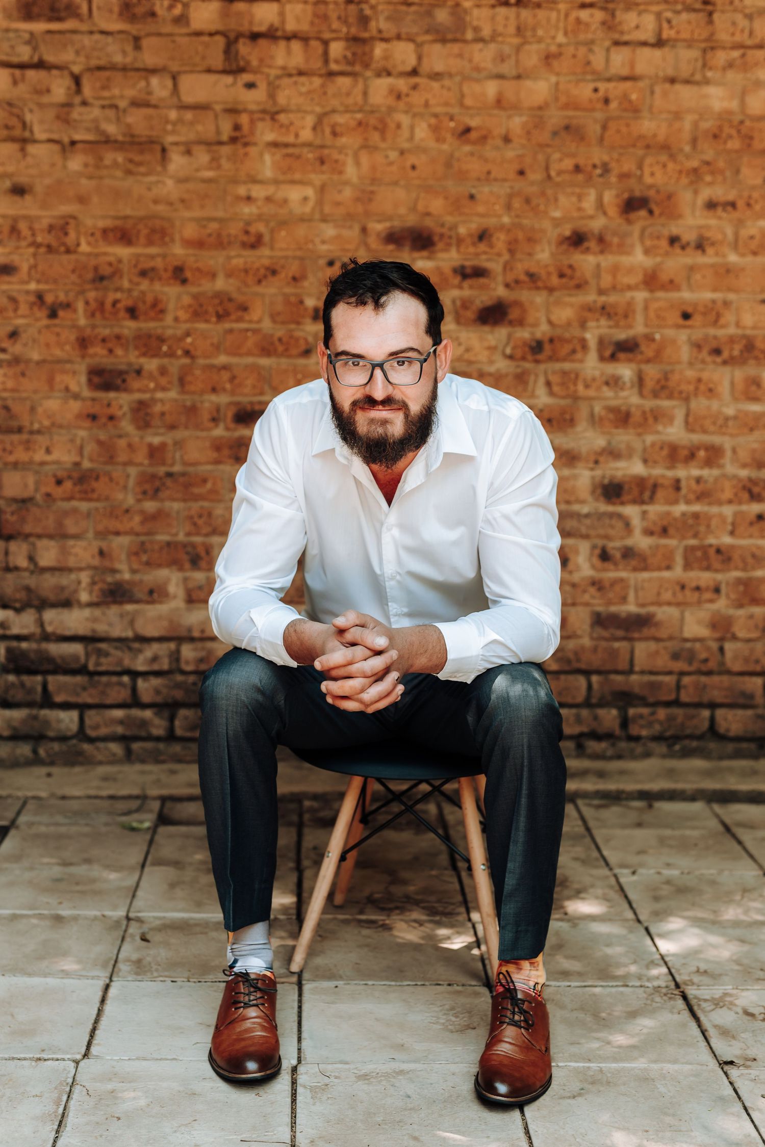 Professional portrait of a bearded person in glasses seated on a chair against a brick wall wearing brown oxford shoes.