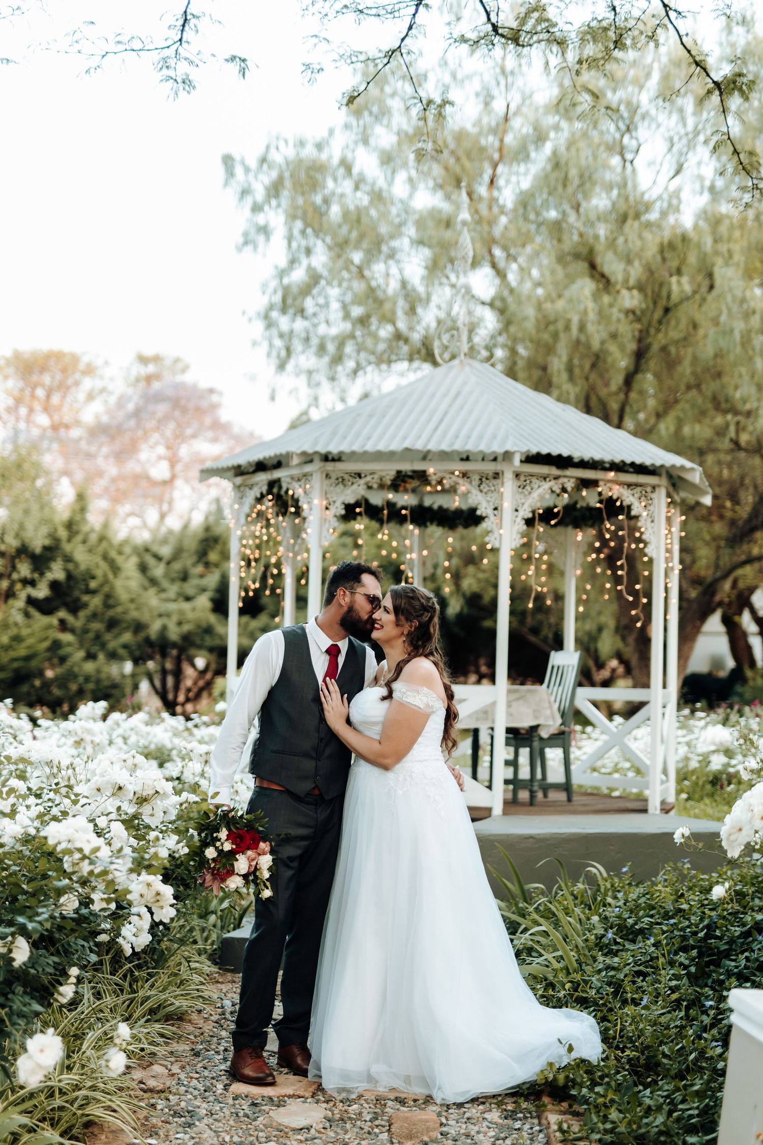 Couple sharing an intimate moment by an ornate white gazebo adorned with twinkling lights.