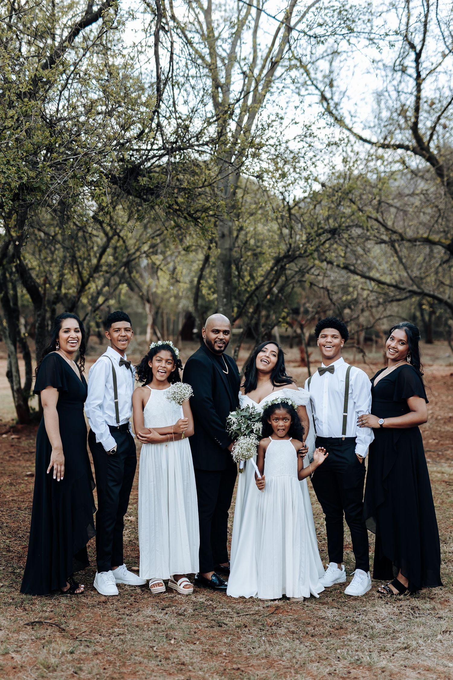 Wedding party in coordinated black and white attire poses together outdoors in a wooded setting.
