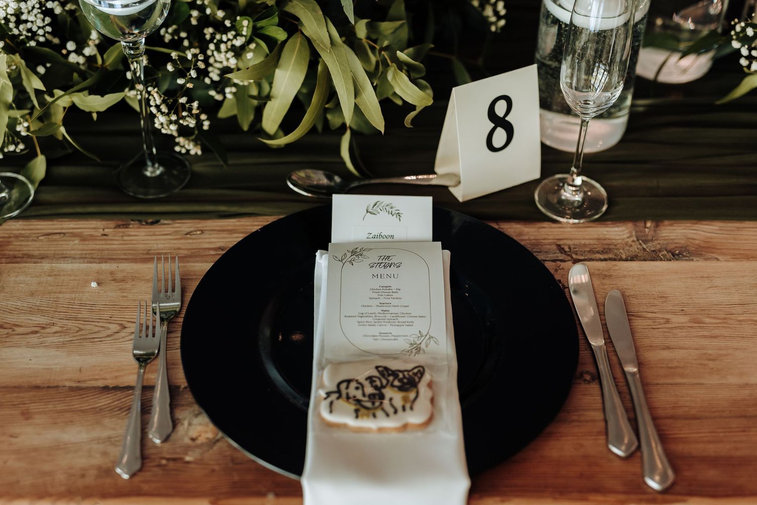 Elegant wedding place setting with black charger plate, menu card and table number 8 on wooden table with greenery.