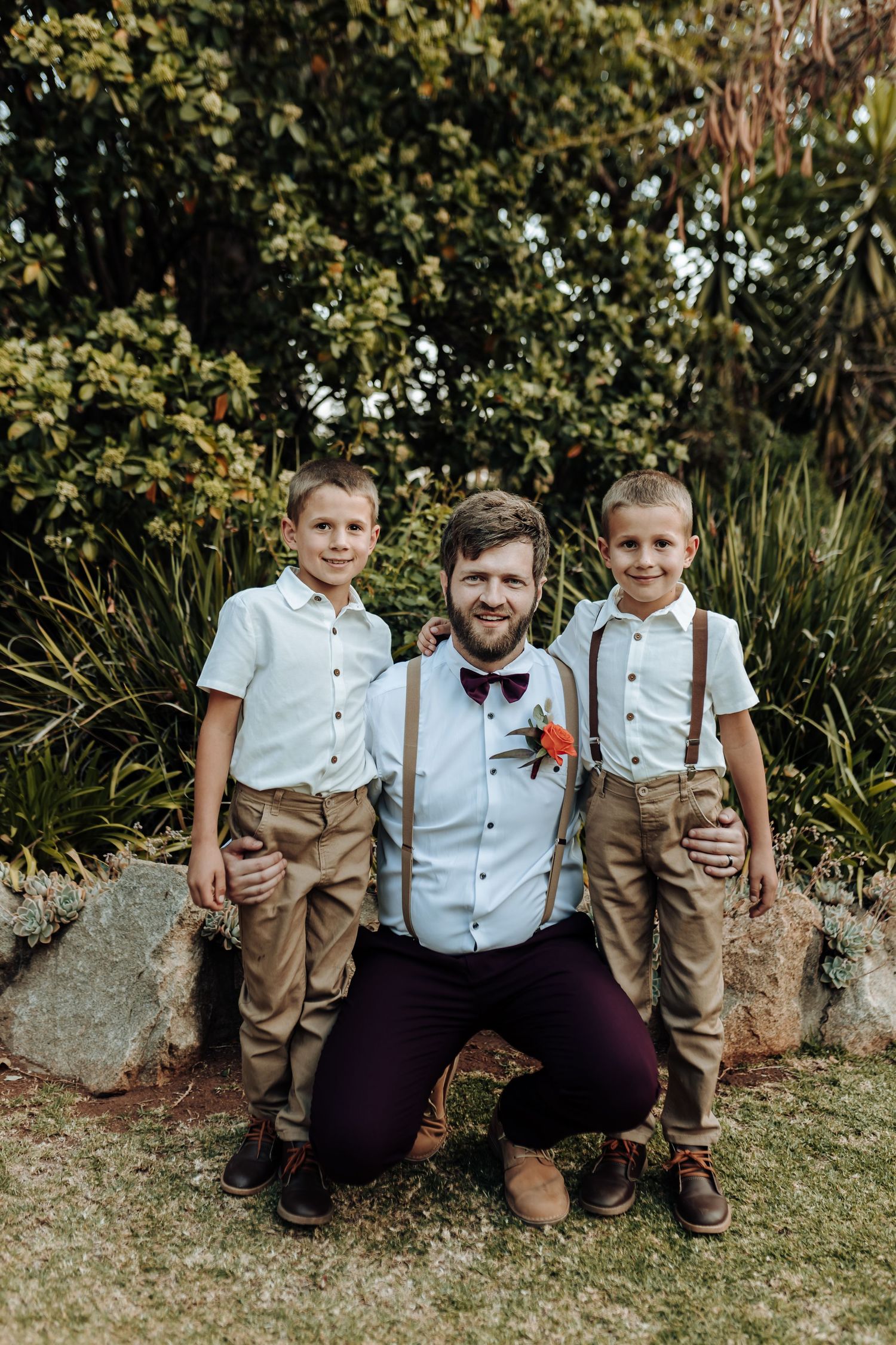 Groom kneeling with young ring bearers in matching white shirts and suspenders.