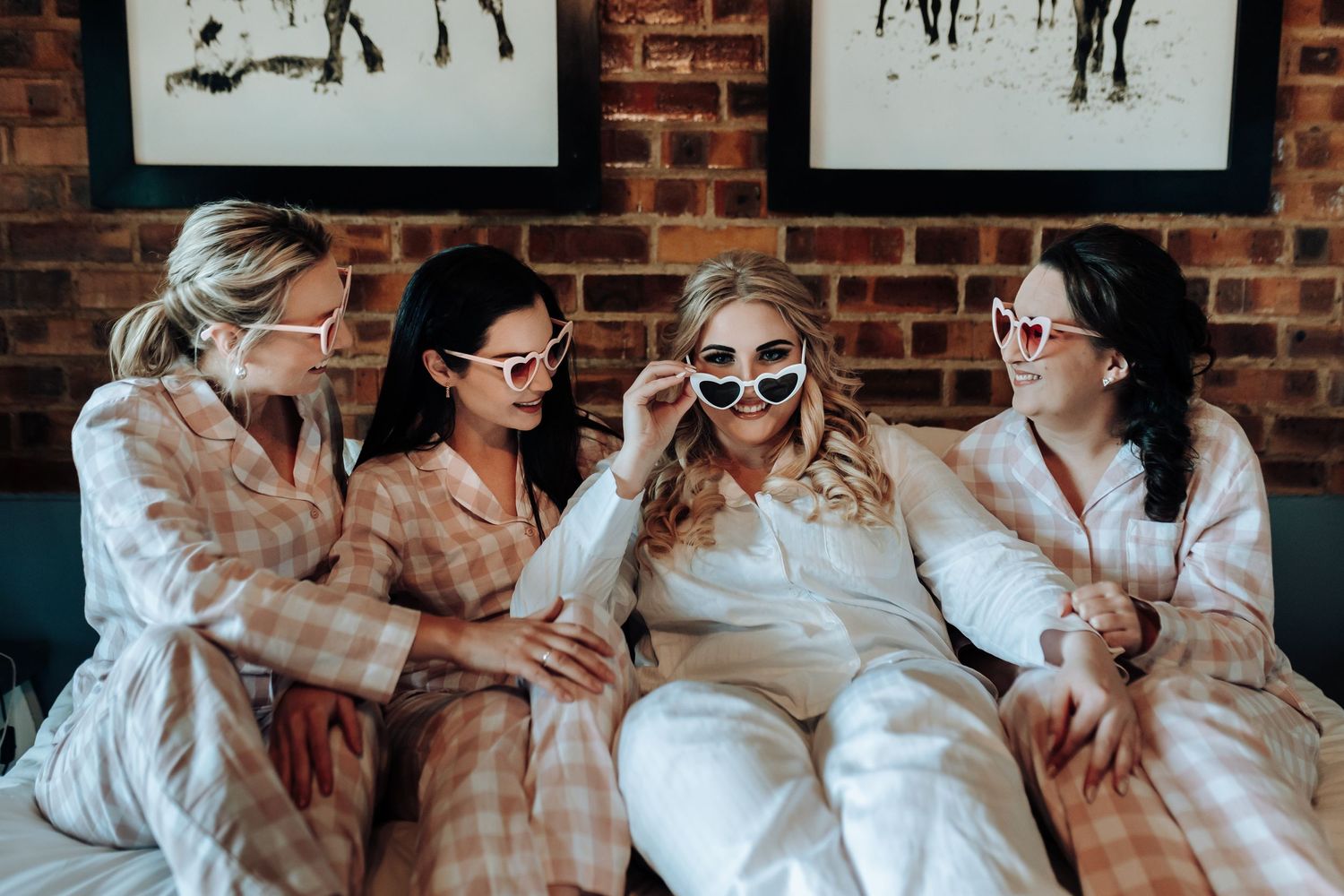 Group in matching pajamas wearing sunglasses pose together on bed with brick wall backdrop.