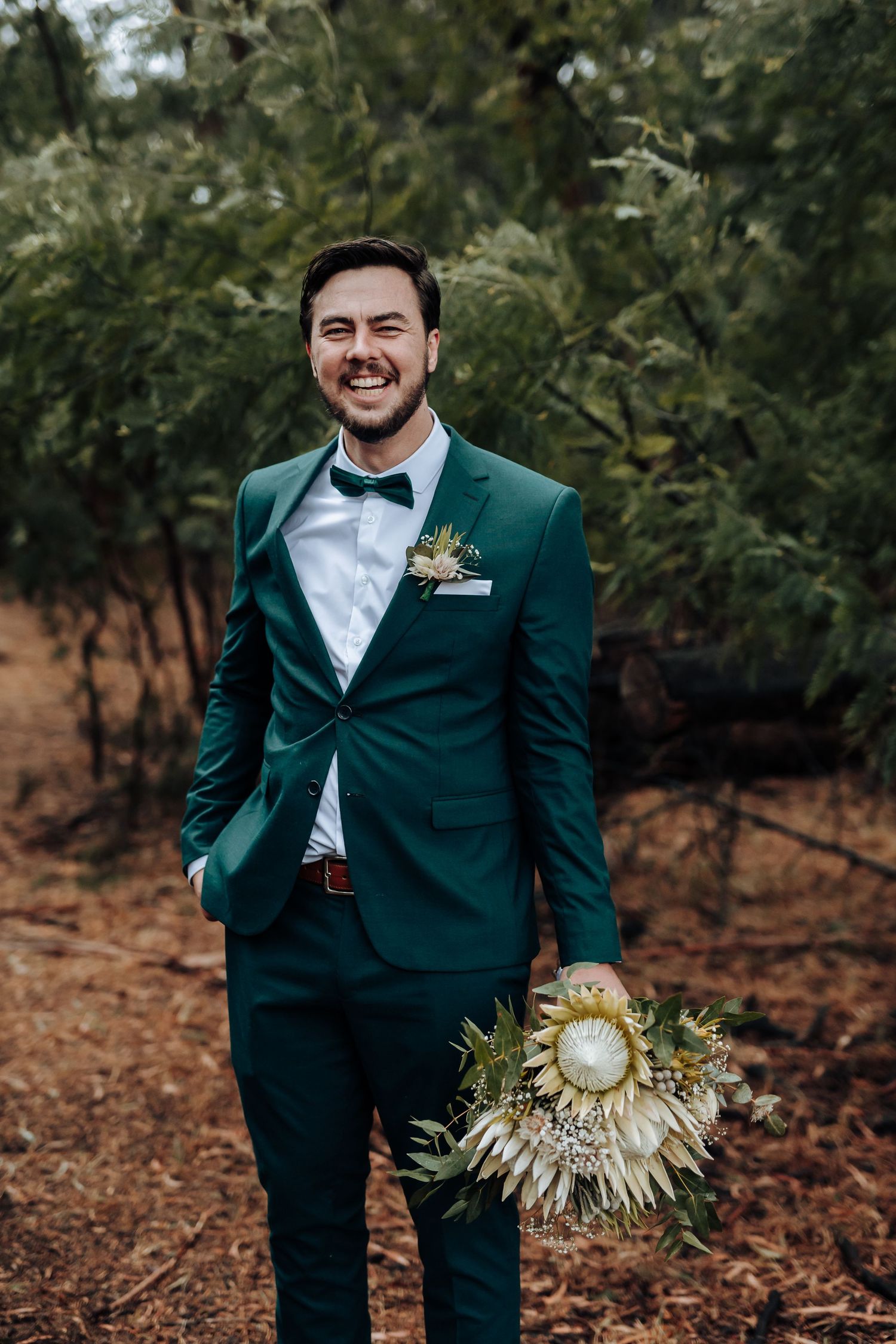 Groom in emerald green suit holding unique dried palm bouquet against forest backdrop.