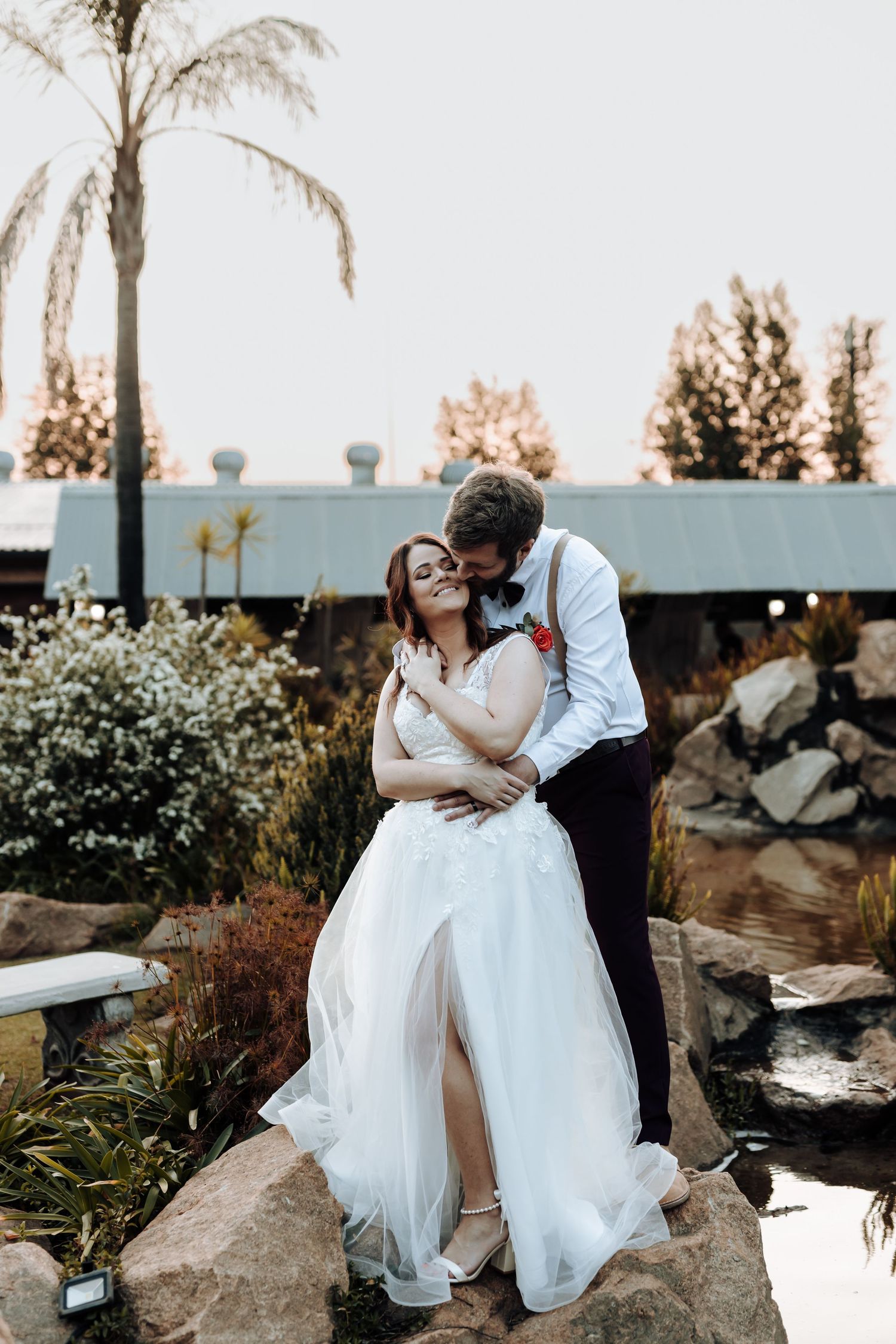 Intimate moment between newlyweds posing on rocky landscape with palm trees in background.