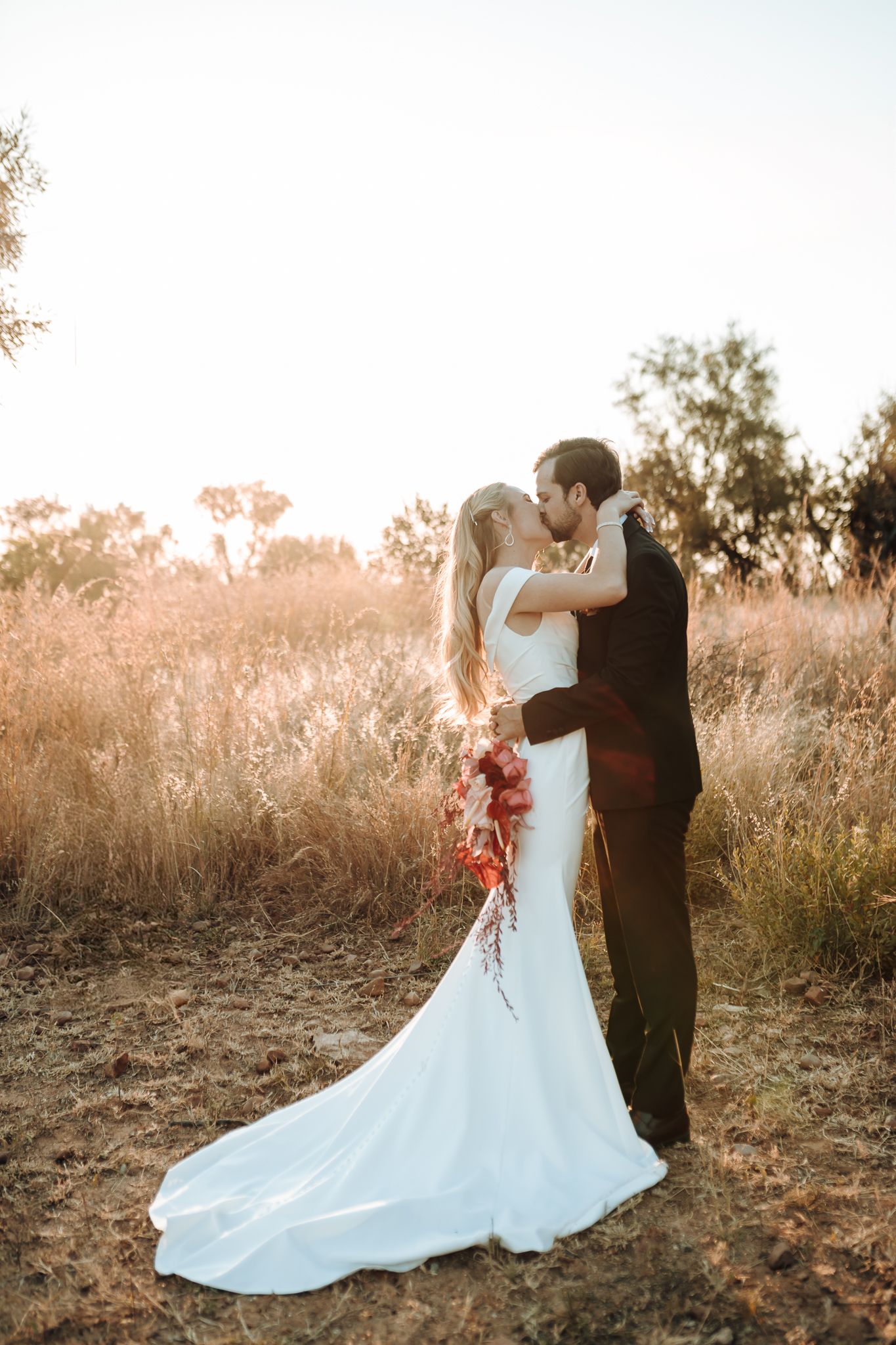 Romantic sunset portrait of newlyweds embracing in golden field with flowing white wedding dress.