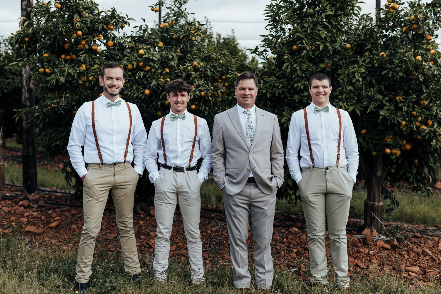 Groomsmen in khaki pants and suspenders pose with groom in gray suit among fruit trees.