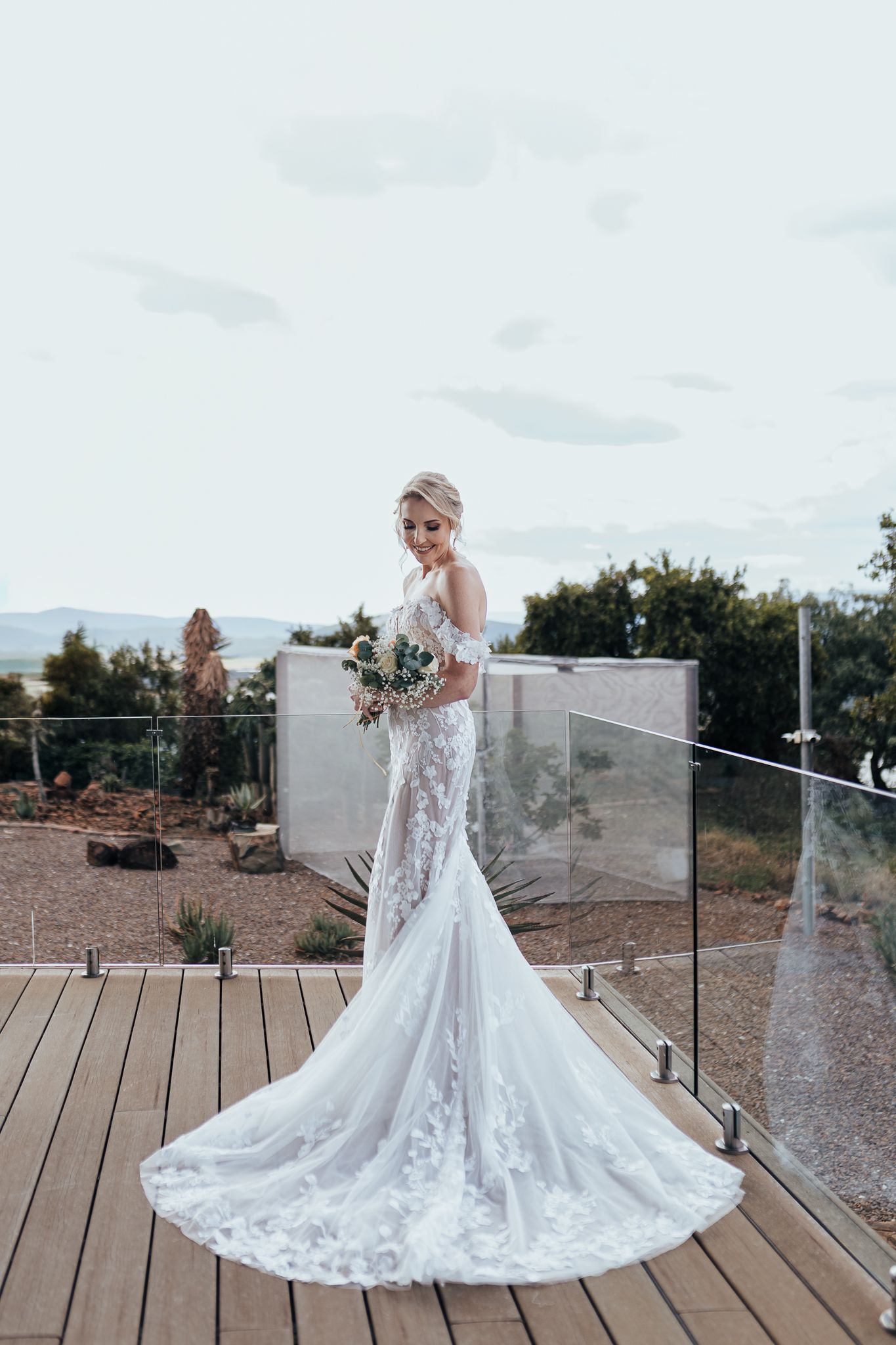 Bride in fitted lace wedding dress poses on deck with mountain landscape backdrop.