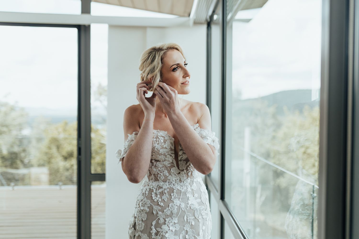 Elegant bridal portrait by floor-to-ceiling windows while putting on earrings.