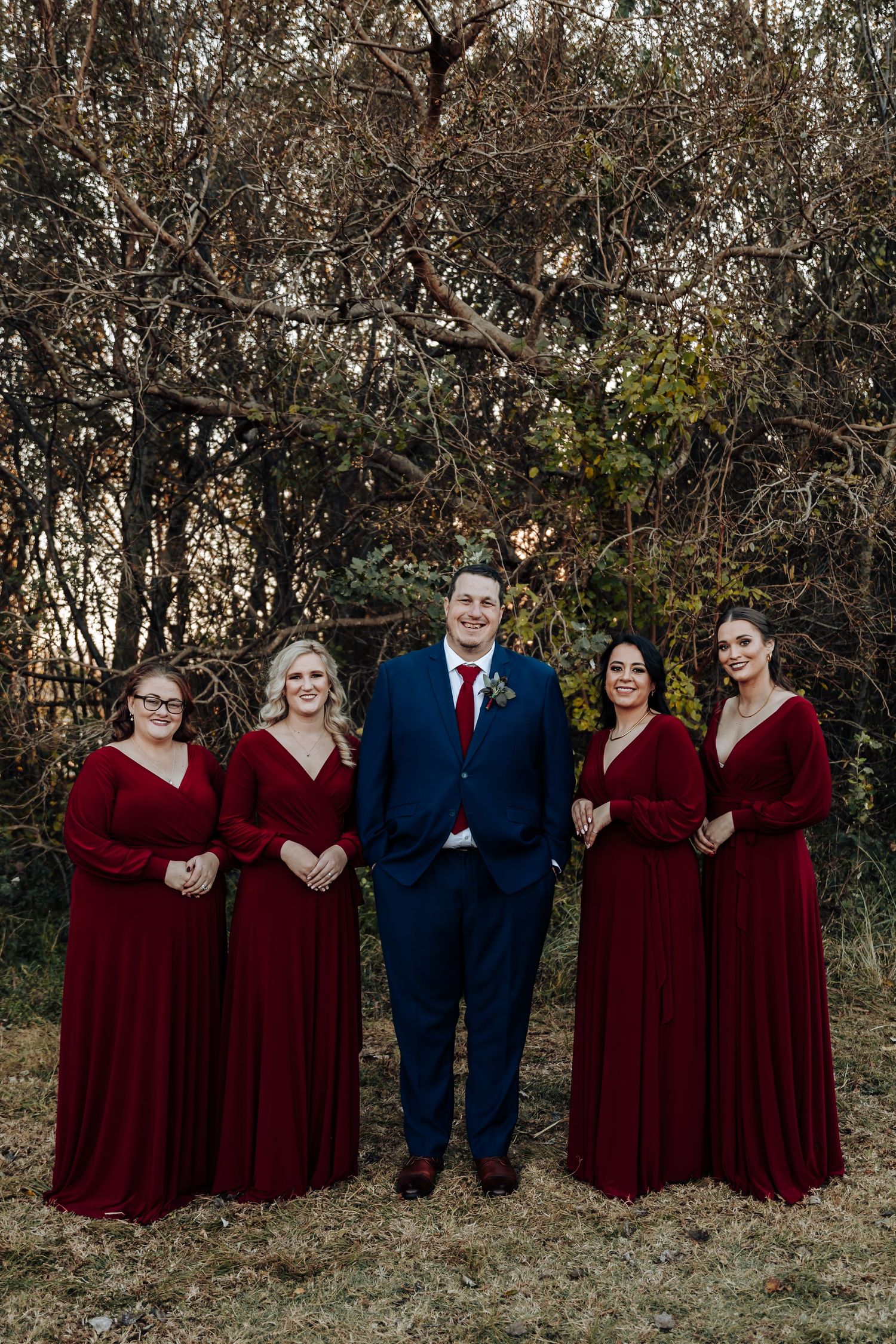 Groom in navy suit with four bridesmaids in burgundy dresses posing outdoors against bare winter trees.