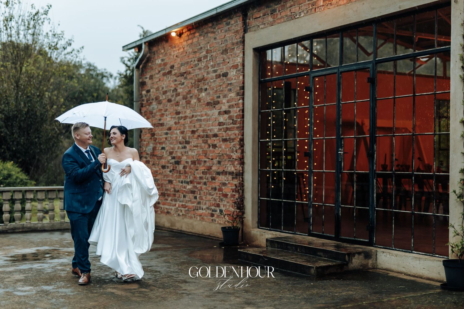 Bride and groom sharing an umbrella outside a rustic brick wedding venue decorated with warm fairy lights.