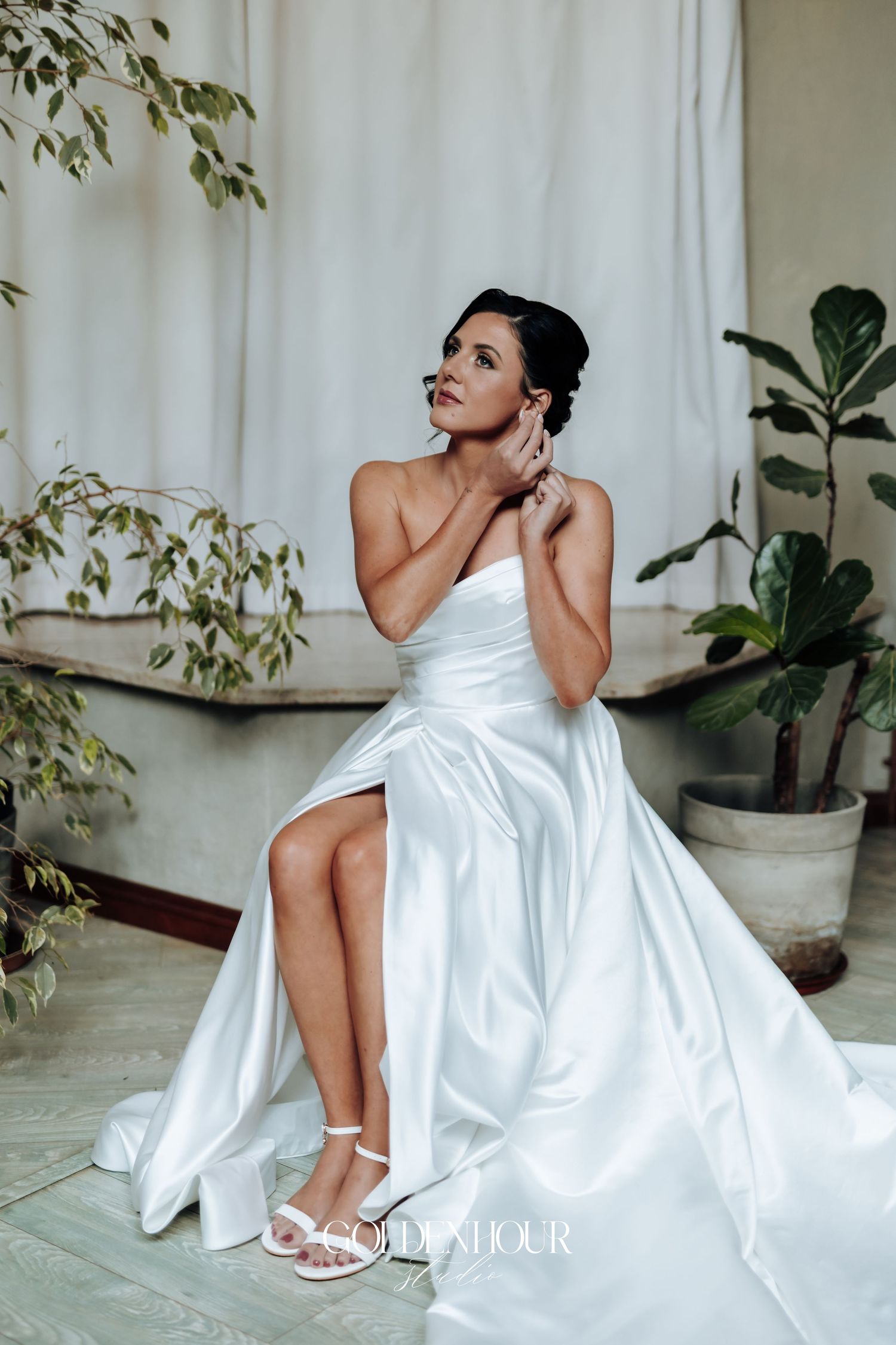 Bride in a white strapless satin wedding gown with a slit, adjusting her earring while seated among green plants.