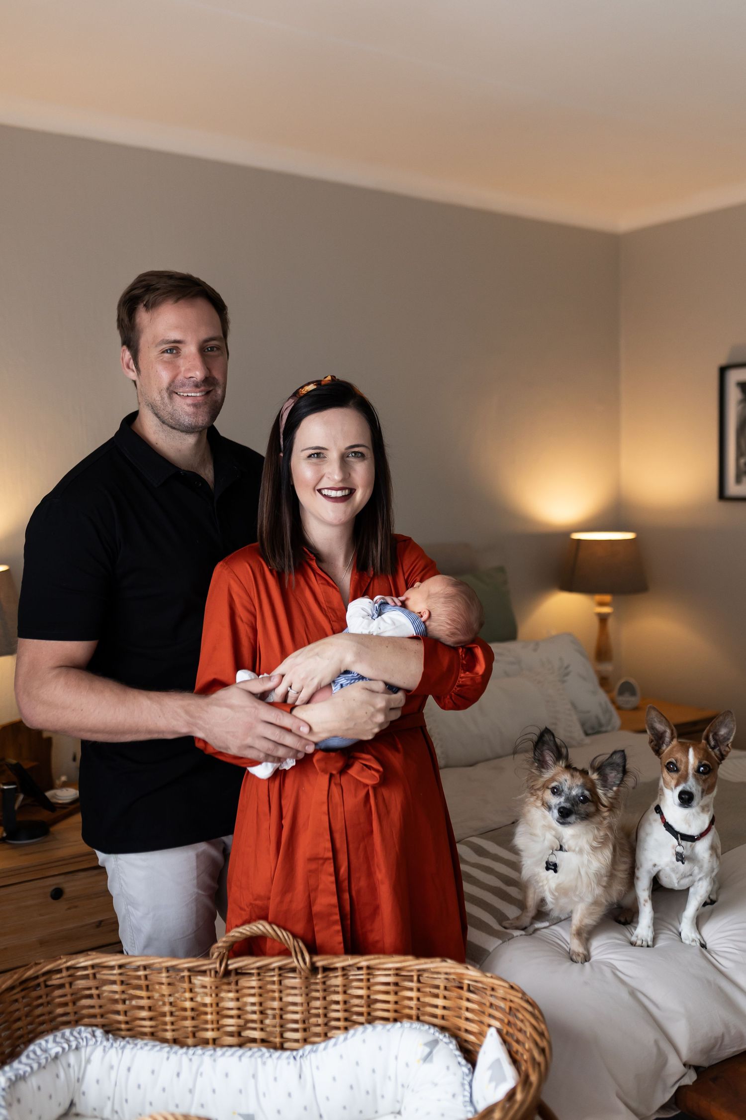 A couple poses with their newborn baby and two dogs in their living room with warm lighting.