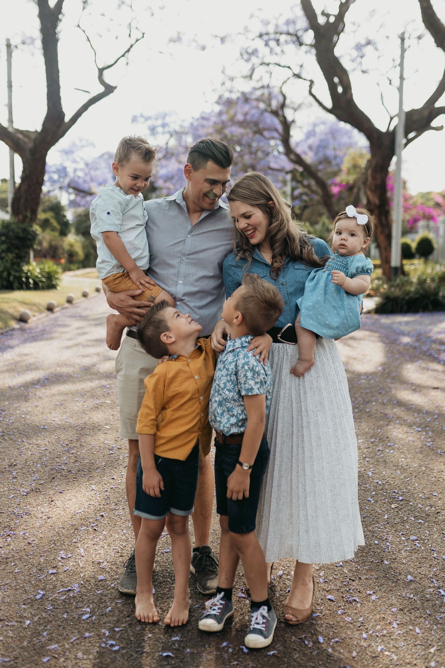 A family shares joyful candid moments together on a path lined with blooming jacaranda trees in the afternoon light.
